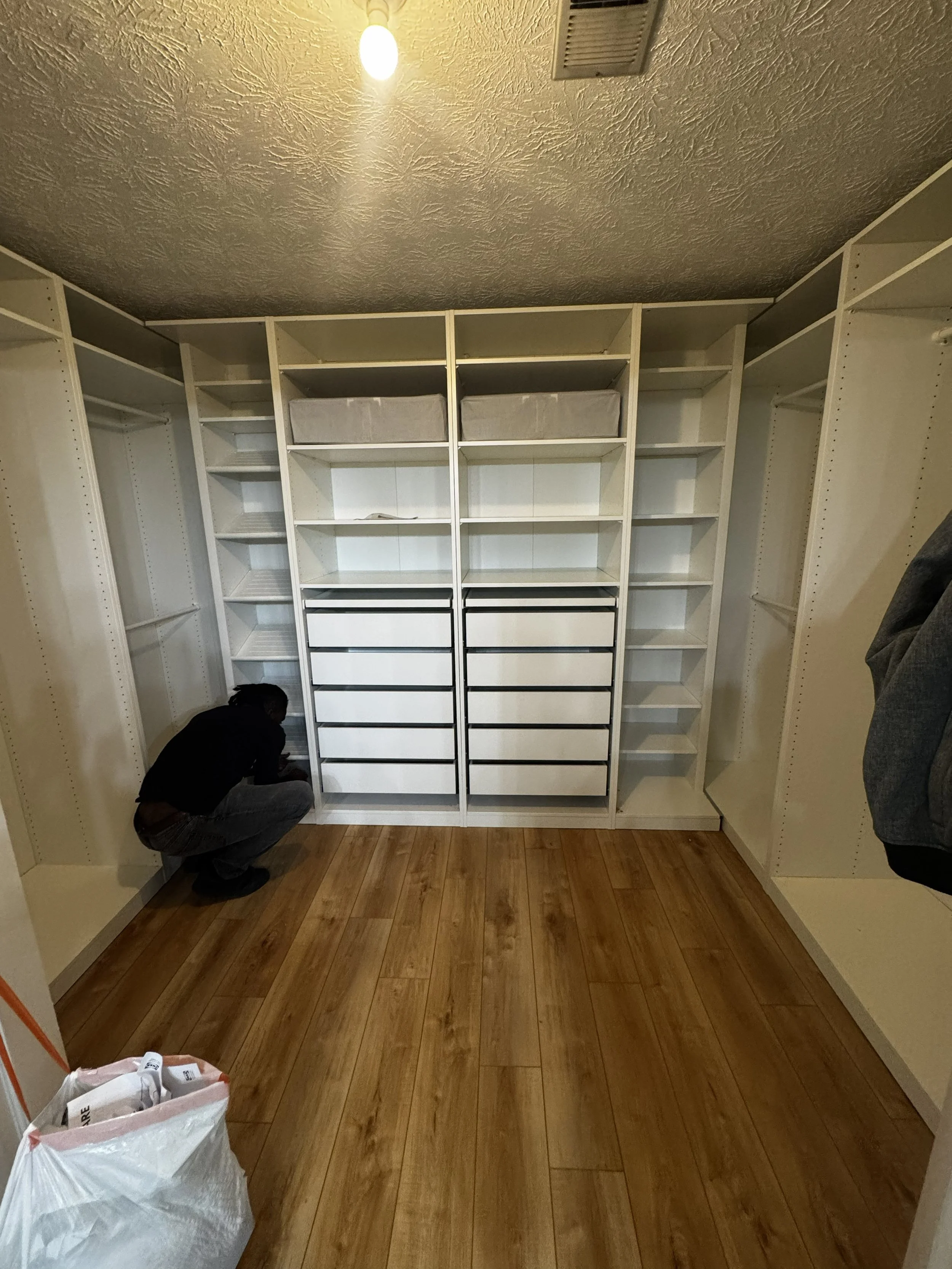 A person squatting and organizing shelves in a walk-in closet with white shelving, drawers, and empty storage compartments, with wooden floor and unfinished wall