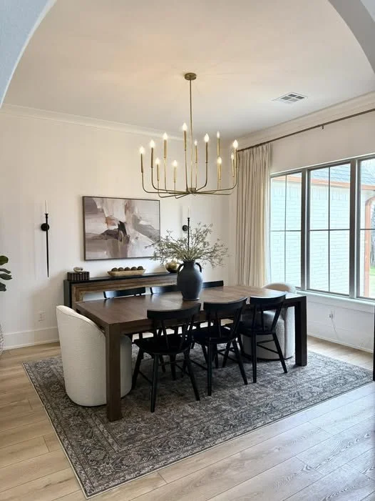 A dining room with a wooden table, six black chairs, a large blue vase with greenery, a chandelier, a wall-mounted artwork, and a window with beige curtains.