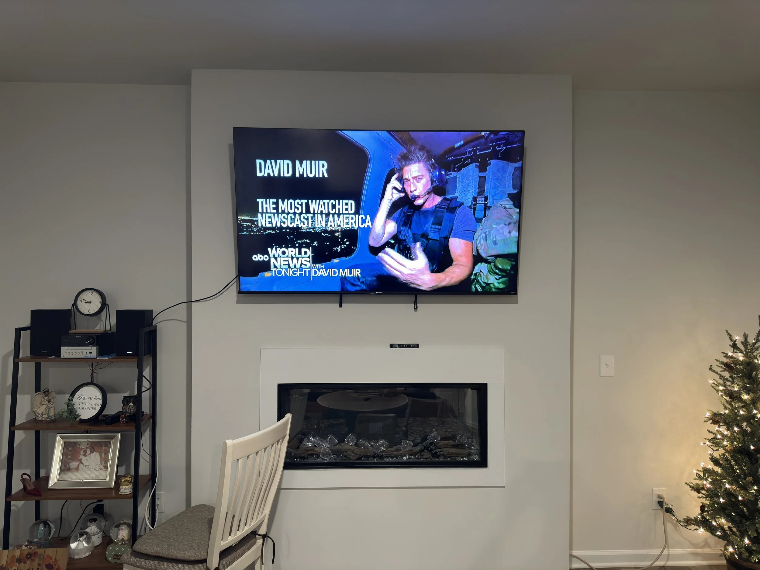 Living room with a mounted TV displaying David Muir on ABC's World News Tonight, a white chair, a black shelf with decorations, a clock, and a Christmas tree with lights.