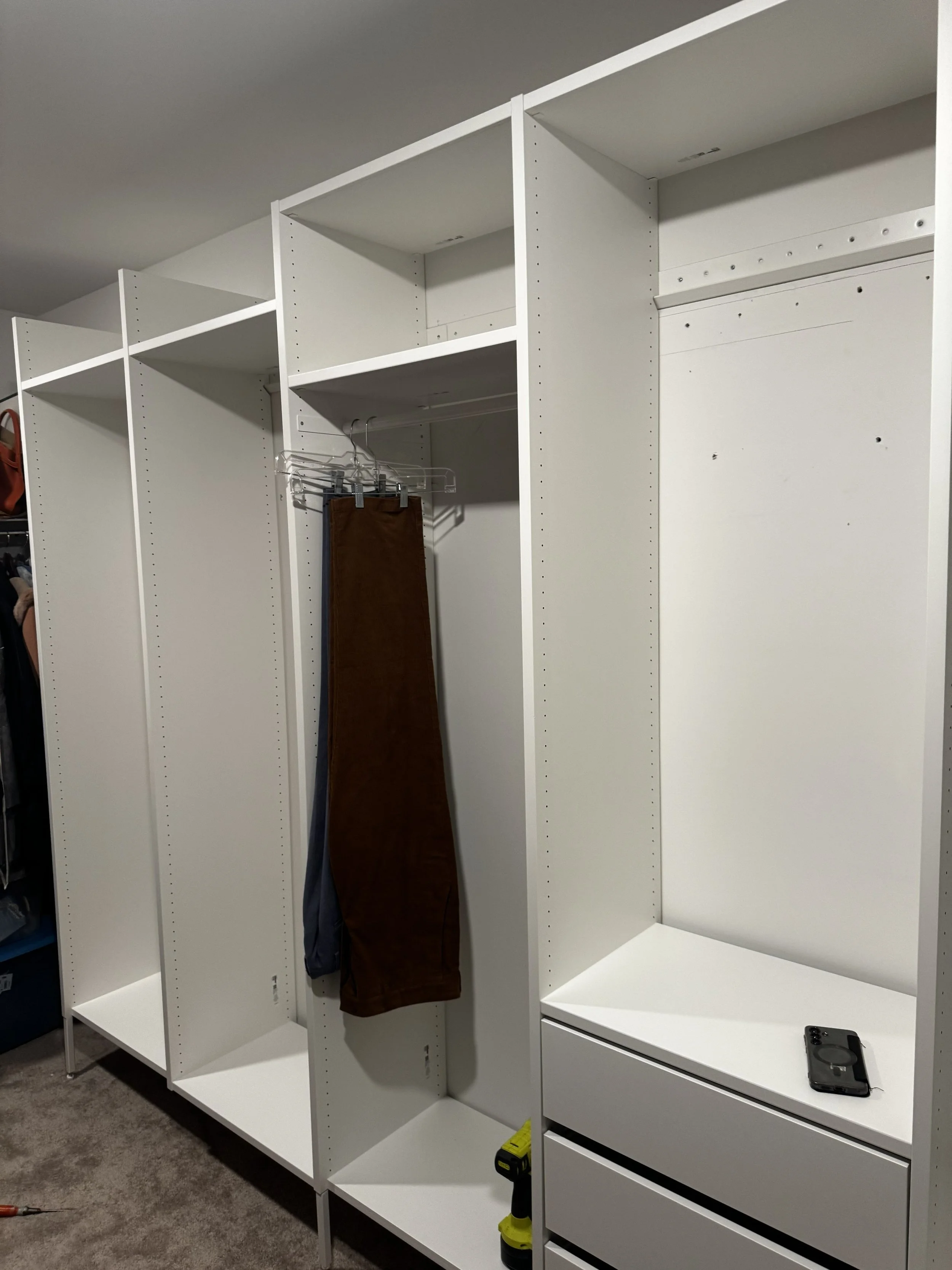Empty white closet with a brown pant and a black shirt hanging on a wire hanger, a yellow tool on the floor, and a black phone on the bottom shelf.