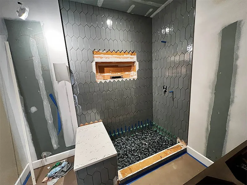 Bathroom with hexagonal dark gray tiles on the shower wall, a small niche with wood framing, and a black pebble shower floor under construction.