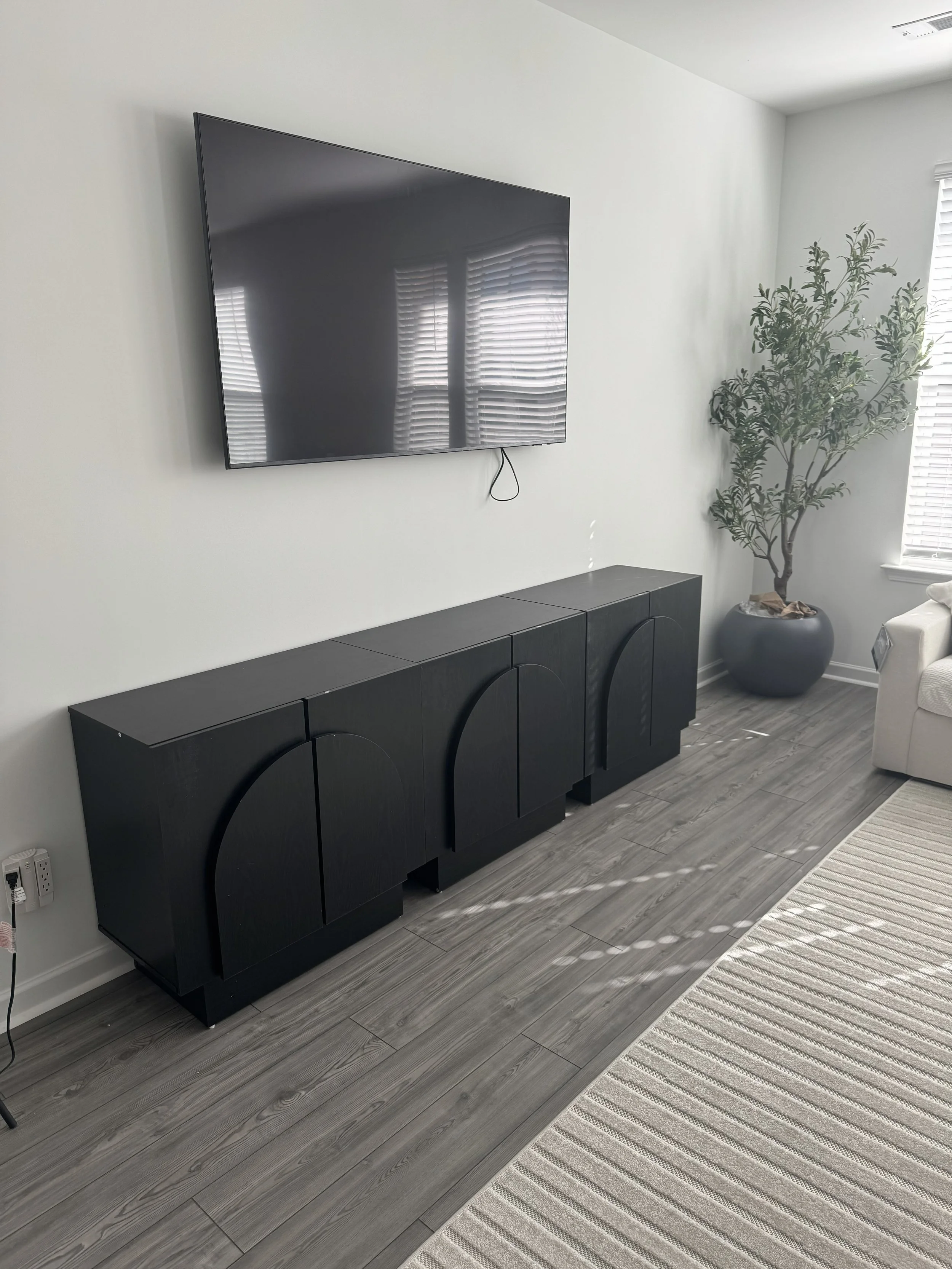 Modern living room with a mounted flat-screen TV, black cabinet, potted plant, window with blinds, and a white armchair.