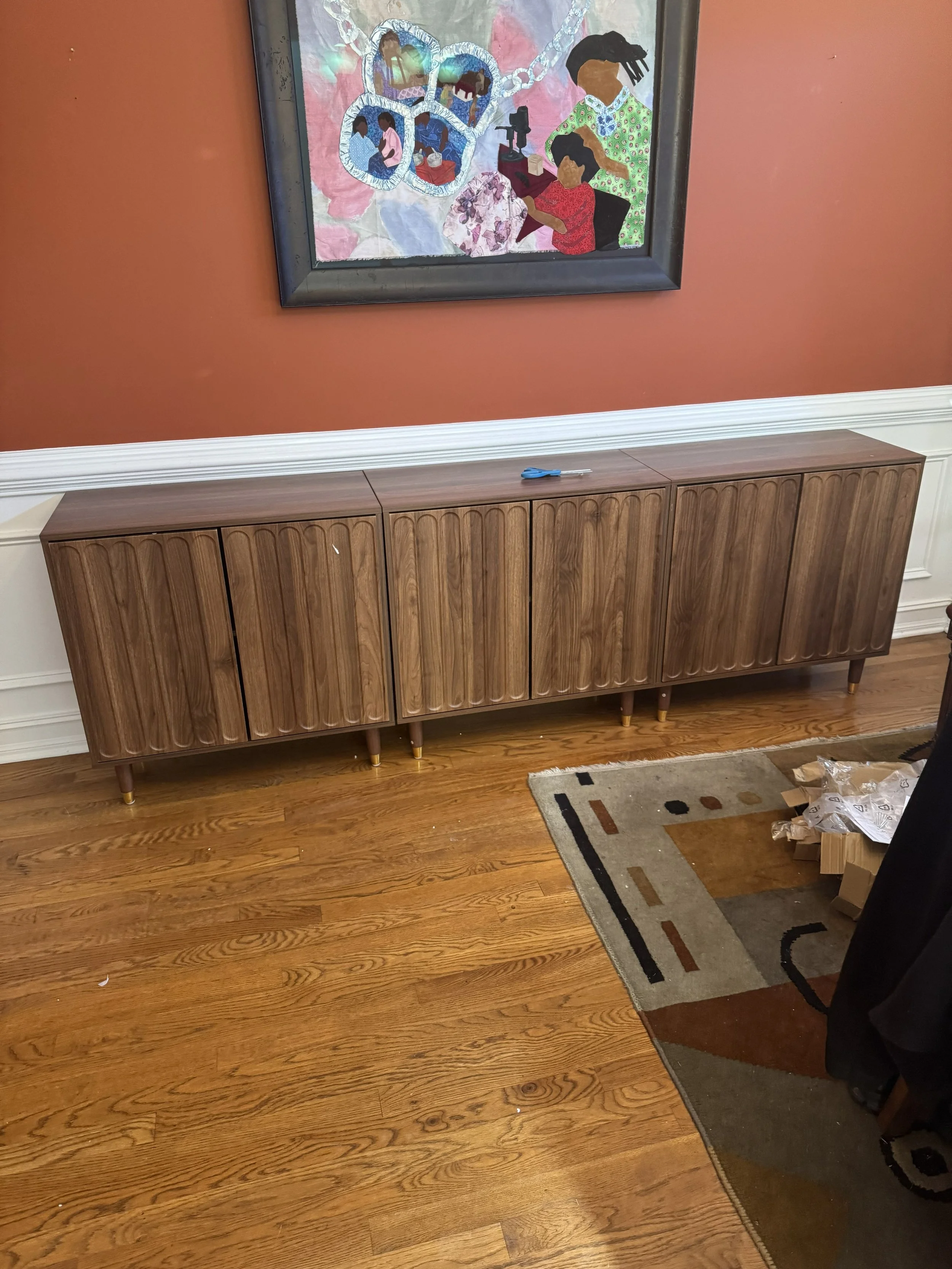 A brown wooden sideboard with a mid-century modern design is placed against a reddish wall with white molding at the bottom. There is a small screwdriver resting on top of the sideboard. Above it, a colorful painting depicting children and people eng