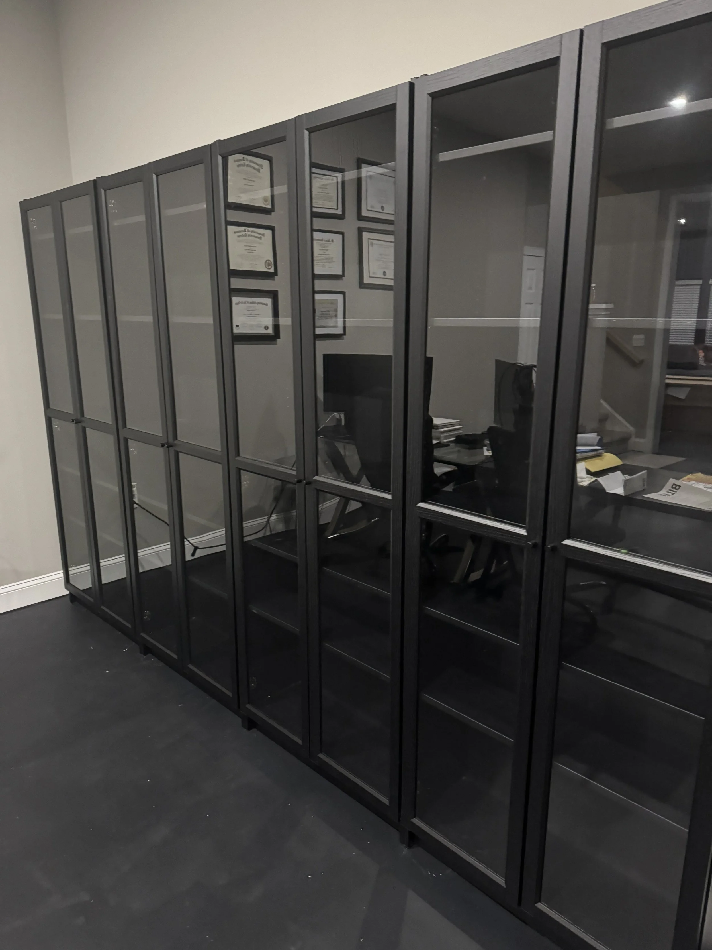 A large black metal and glass cabinet with multiple compartments, containing some office certificates or diplomas on the wall behind it, and an office desk with a computer and scattered papers inside.