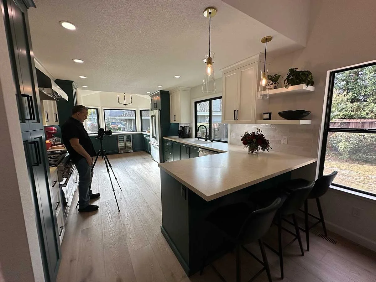 A person setting up a camera in a modern kitchen with green and white cabinets, wooden flooring, pendant lights, and large windows showing an outdoor yard.