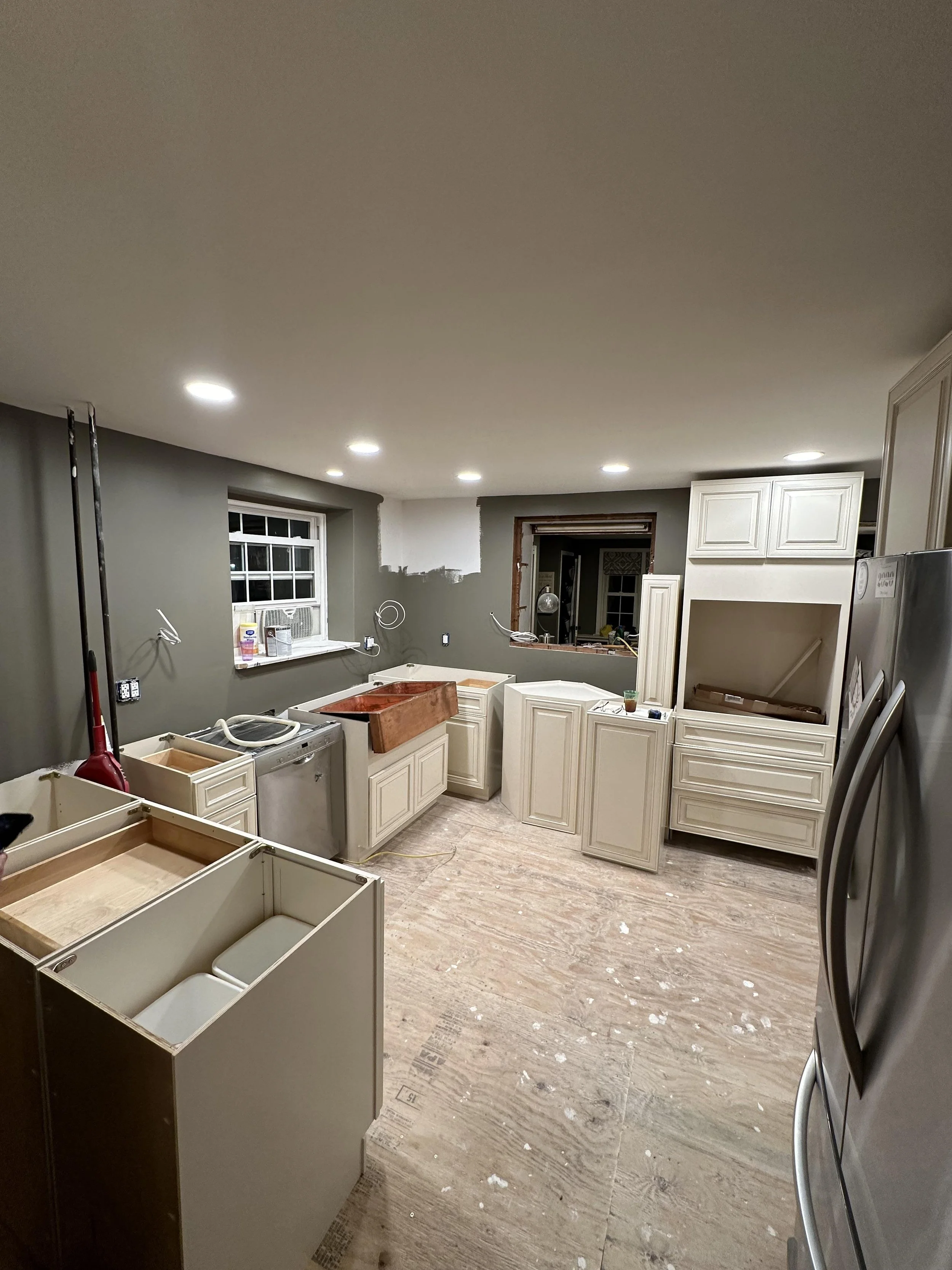 Kitchen under renovation with unfinished cabinets, a copper sink, construction materials, and tools, with a window and recessed ceiling lights.