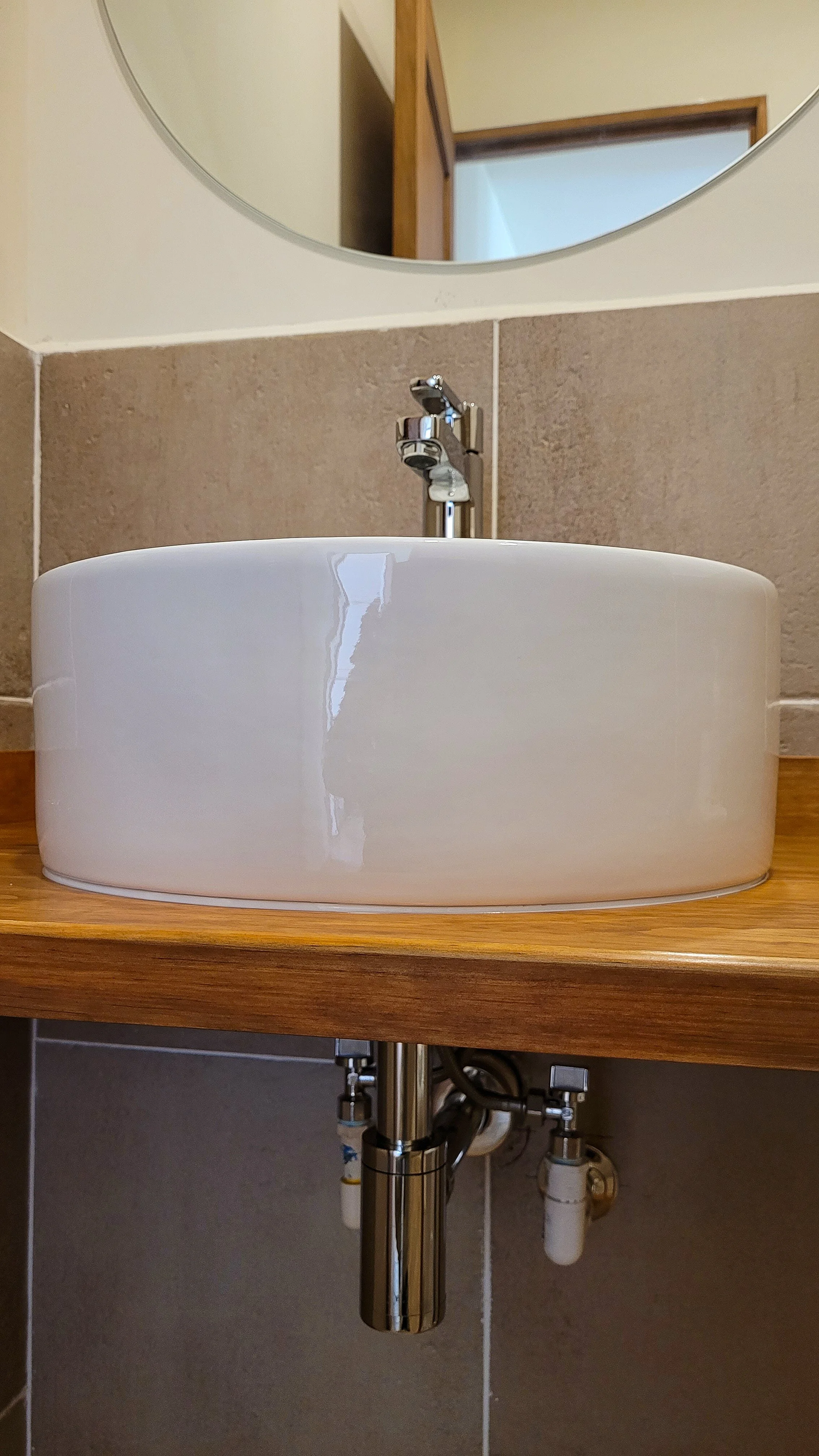 White vessel sink with a chrome single-handle faucet on a wood vanity, with a chrome bottle trap and angle stop valves below.