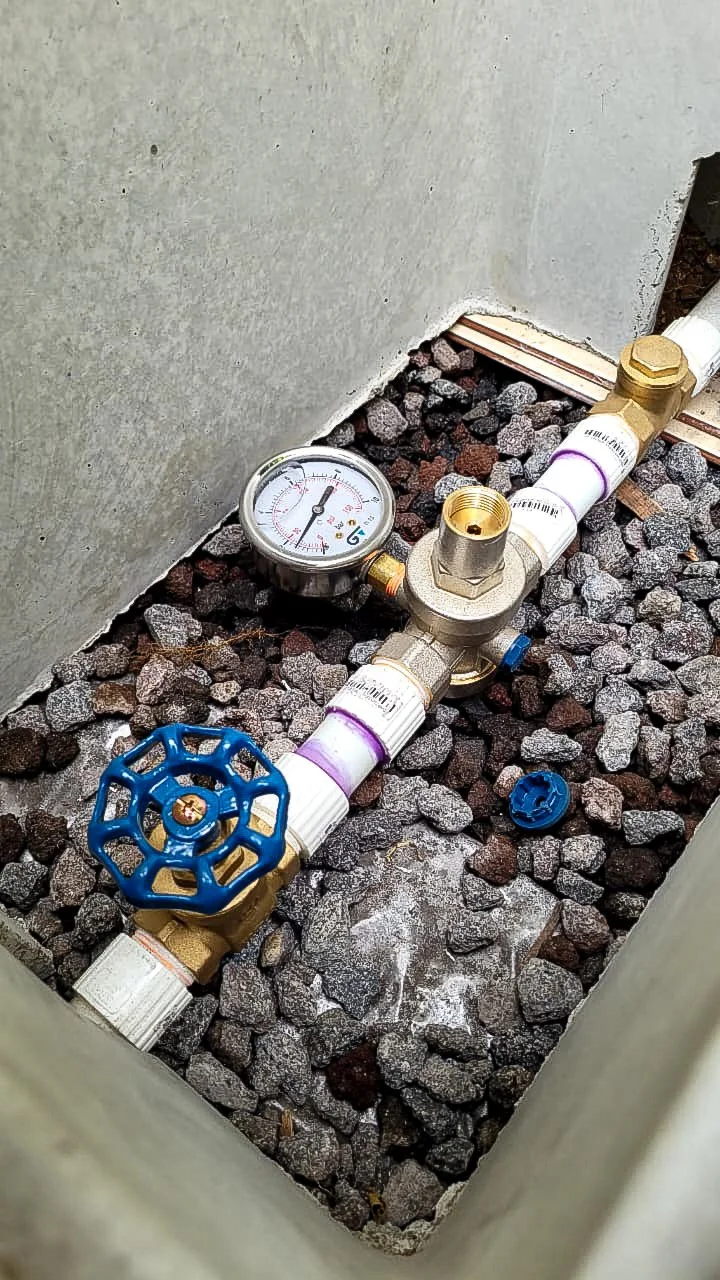 Brass water pressure reducing valve with a gauge and a gate shutoff valve, on PVC line in a concrete box over volcanic gravel.