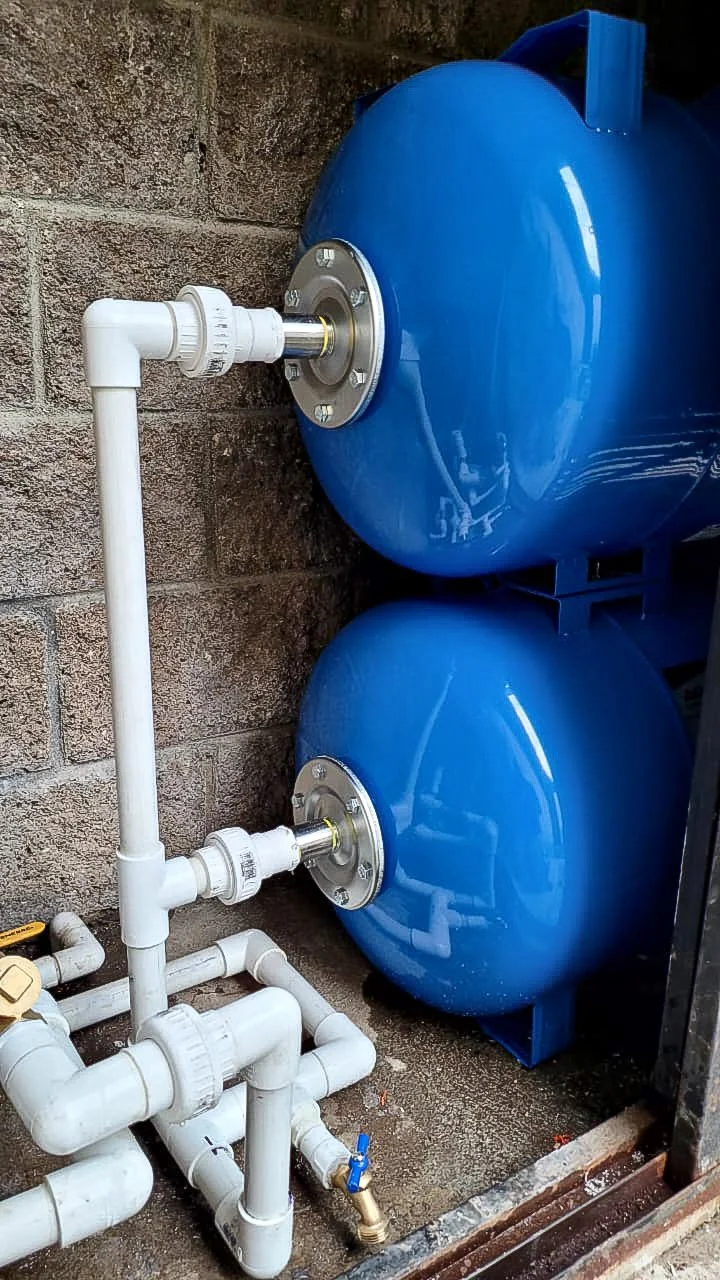 Two stacked blue horizontal pressure tanks connected in parallel with white PVC piping, unions, and a drain valve.