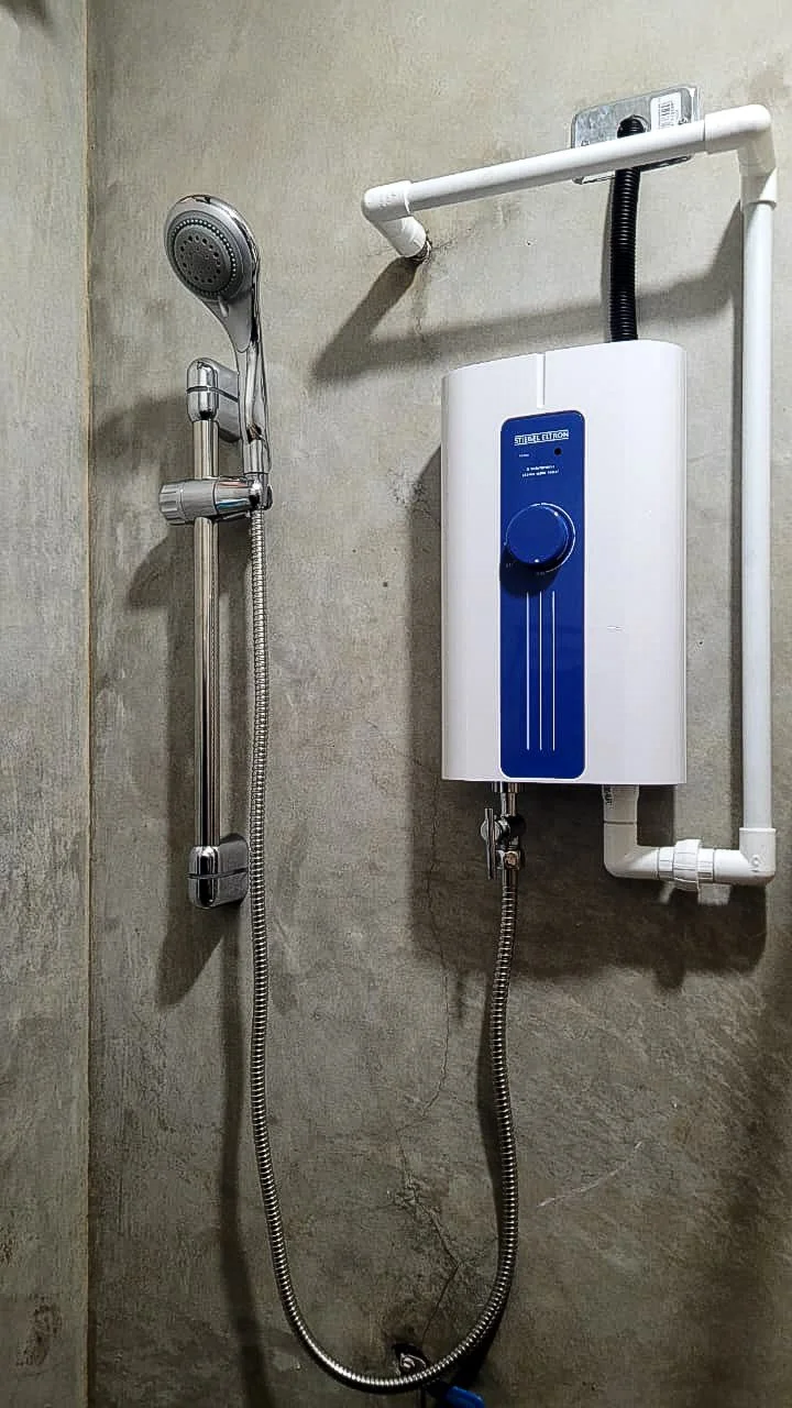 Stiebel Eltron tankless electric shower heater with a blue control dial, mounted on a concrete wall and fed by white PVC piping with a union. It connects to a chrome slide-bar handheld showerhead with a stainless steel hose.