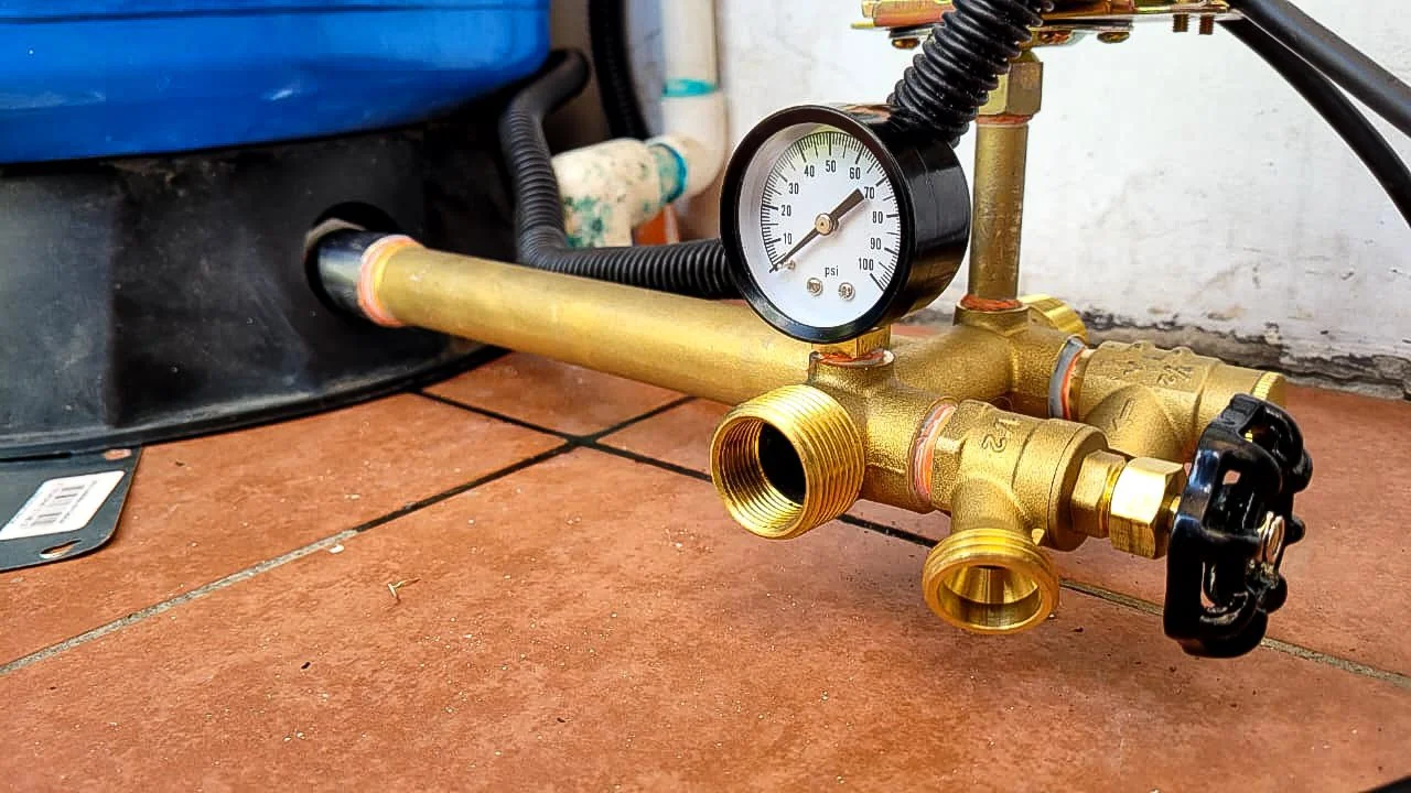 Brass pressure tank tee with a 0–100 psi pressure gauge, pressure switch, and drain valve, connected to a blue well pressure tank during a water system installation.
