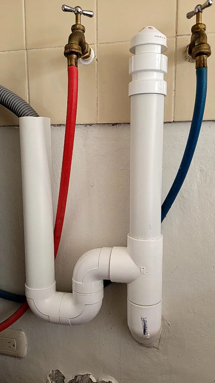 Washing machine drain made of white PVC: a standpipe holding the gray drain hose, a P-trap, and a sanitary tee rising to an air admittance valve. Brass hot and cold faucets feed red and blue supply hoses on a tiled wall.