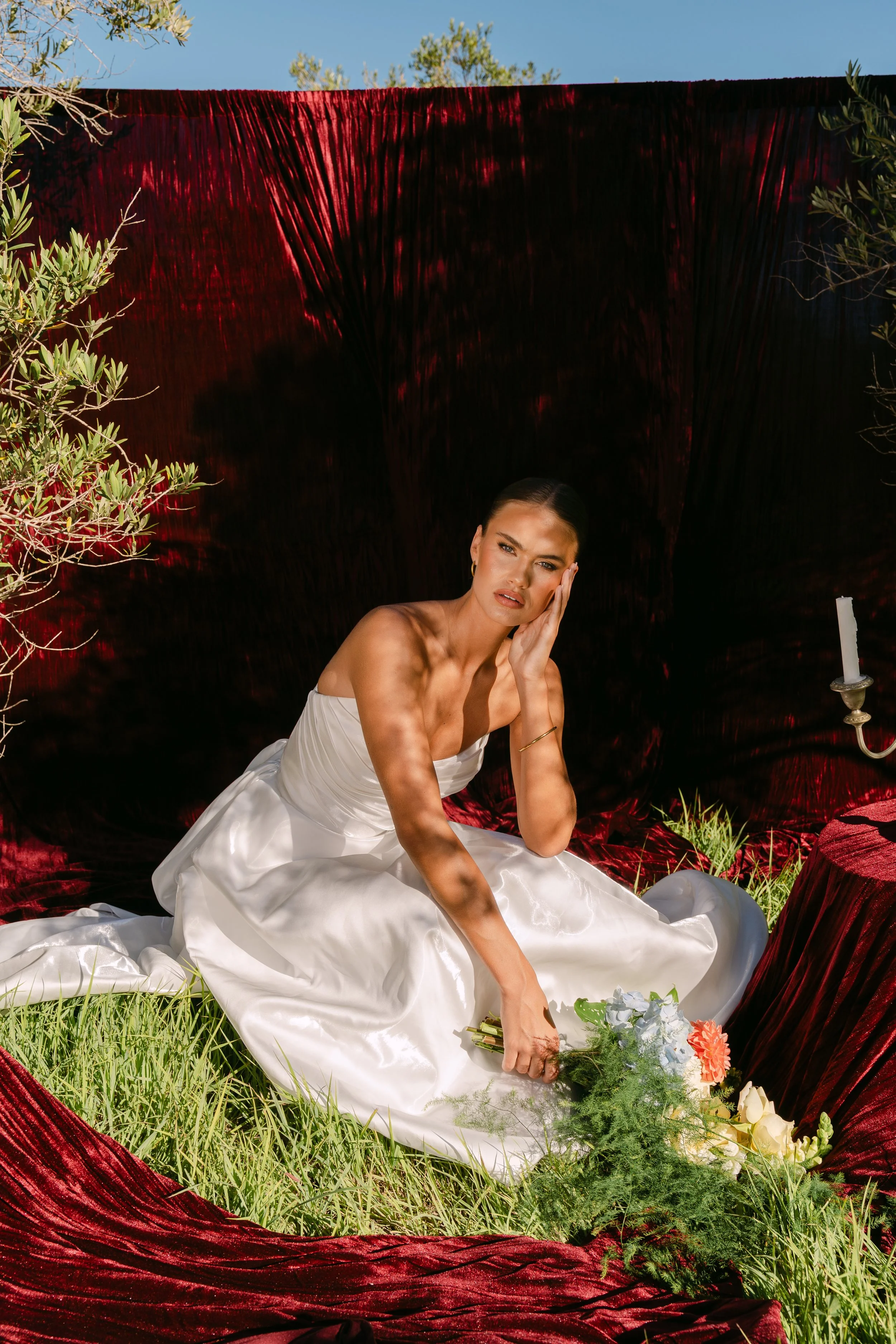 A woman in a white satin wedding dress sitting on the grass, holding a bouquet of flowers, with her hand on her face, surrounded by red velvet fabric and greenery.