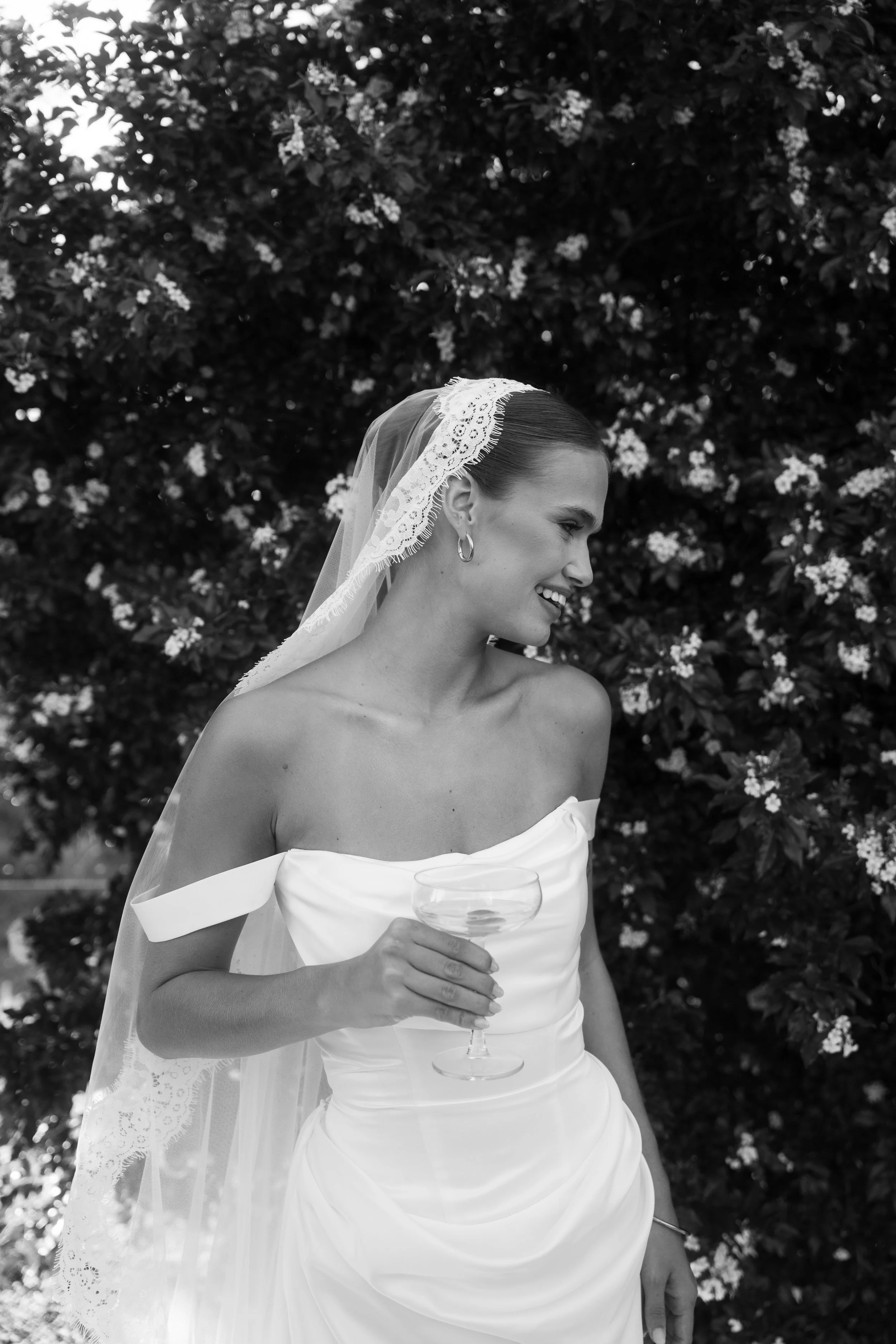 A bride with a lace veil and earrings, smiling and holding a glass of champagne outdoors with a flowered bush in the background.