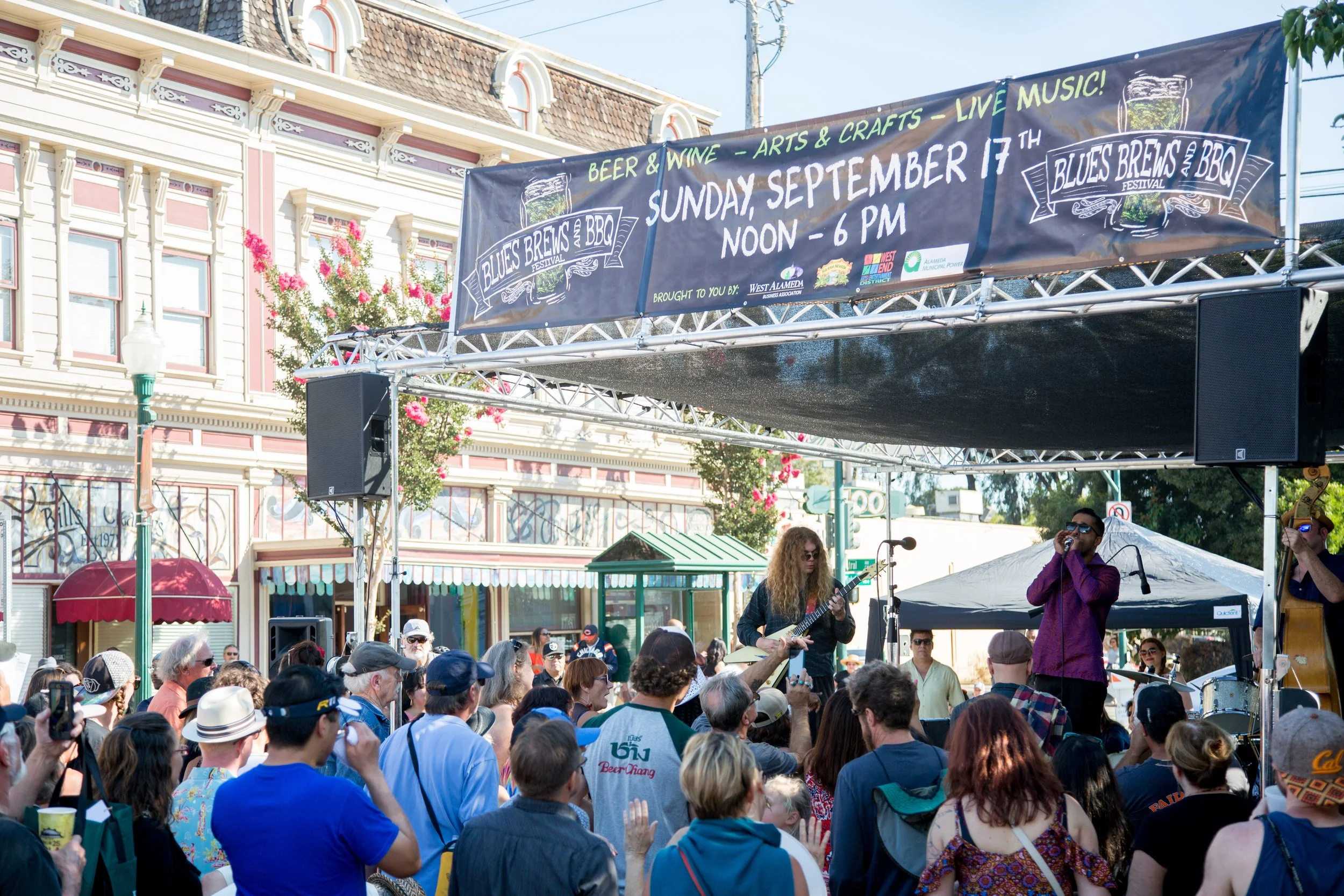 Outdoor festival scene with a stage featuring a live band, a banner reading 'Blues Brews and BBQ Festival' scheduled for September 17th, and a crowd of attendees enjoying the music.