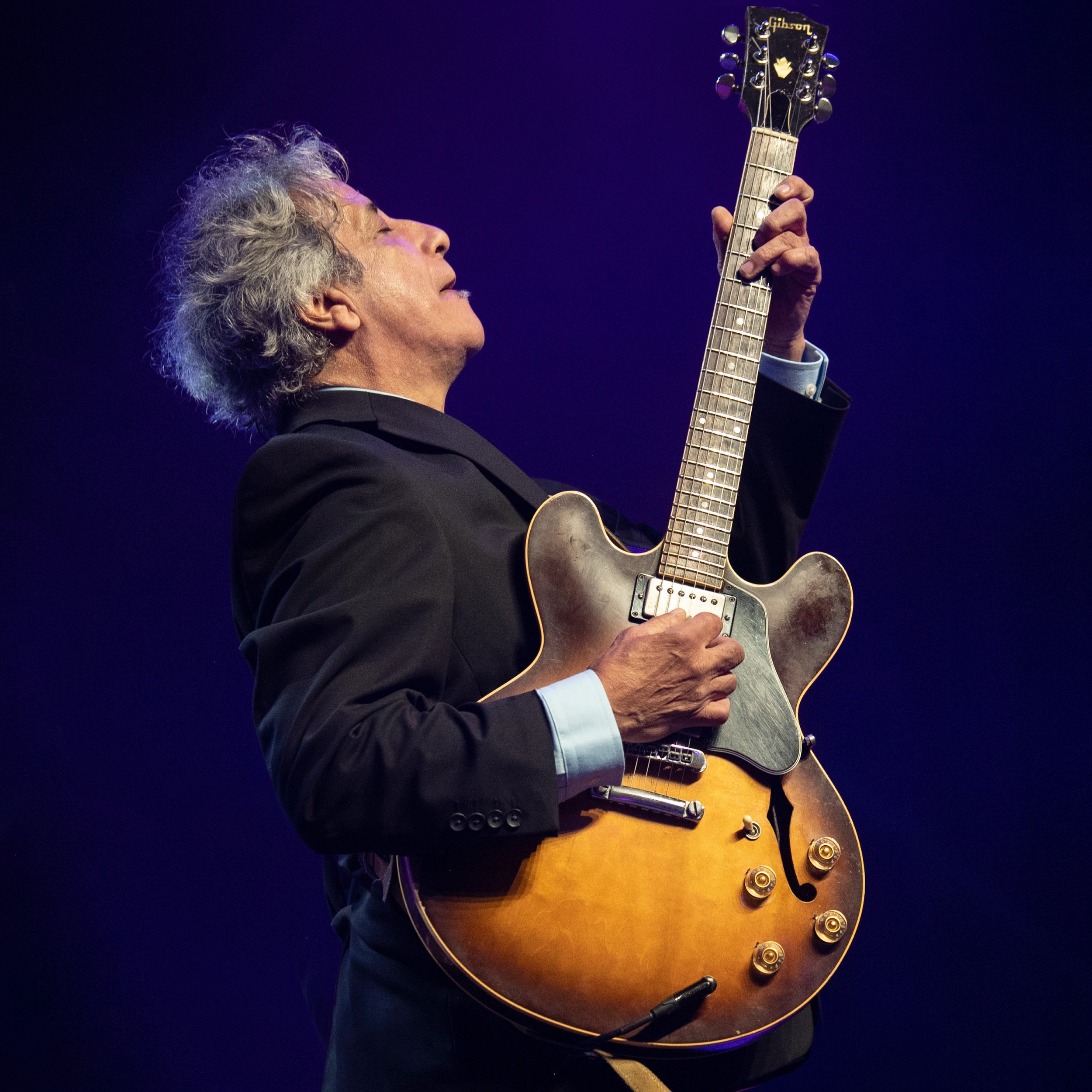 A man with gray curly hair playing an acoustic Gibson guitar on stage.