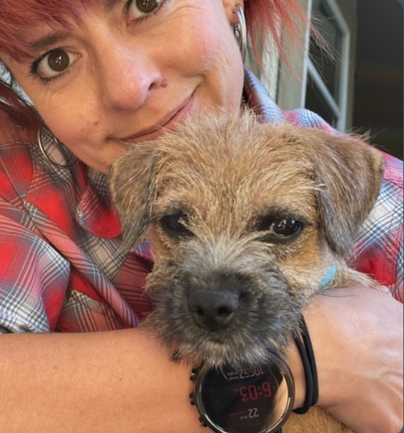 A woman with short pink hair and hoop earrings holding a brown and gray dog close to her face. The woman is smiling slightly, wearing a red plaid shirt, and the dog has a scruffy coat and looks at the camera. The woman is wearing a black watch with a red digital display.