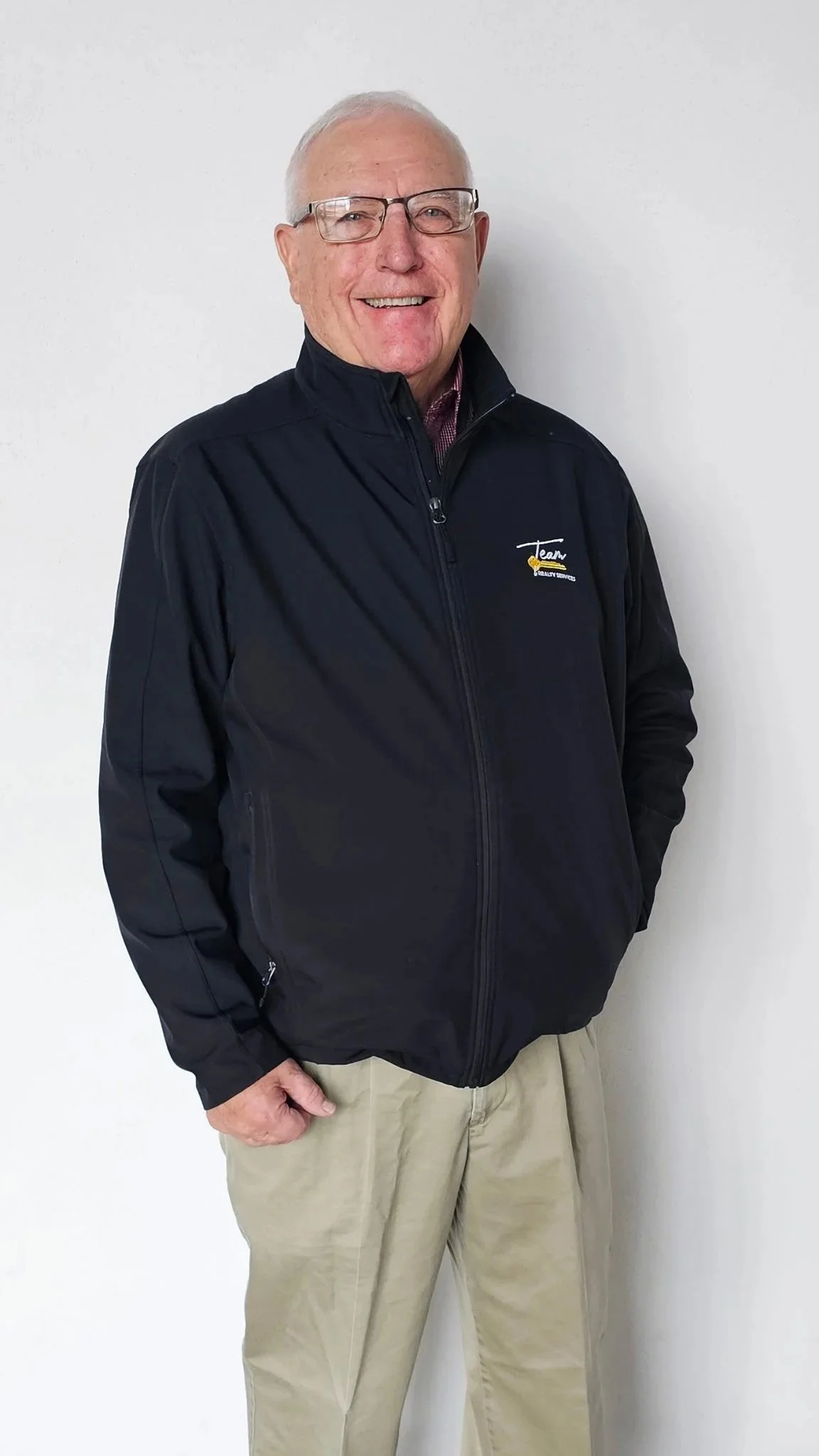 An older man with gray hair and glasses smiling, wearing a black zip-up jacket with a logo on the left chest and standing against a white wall.