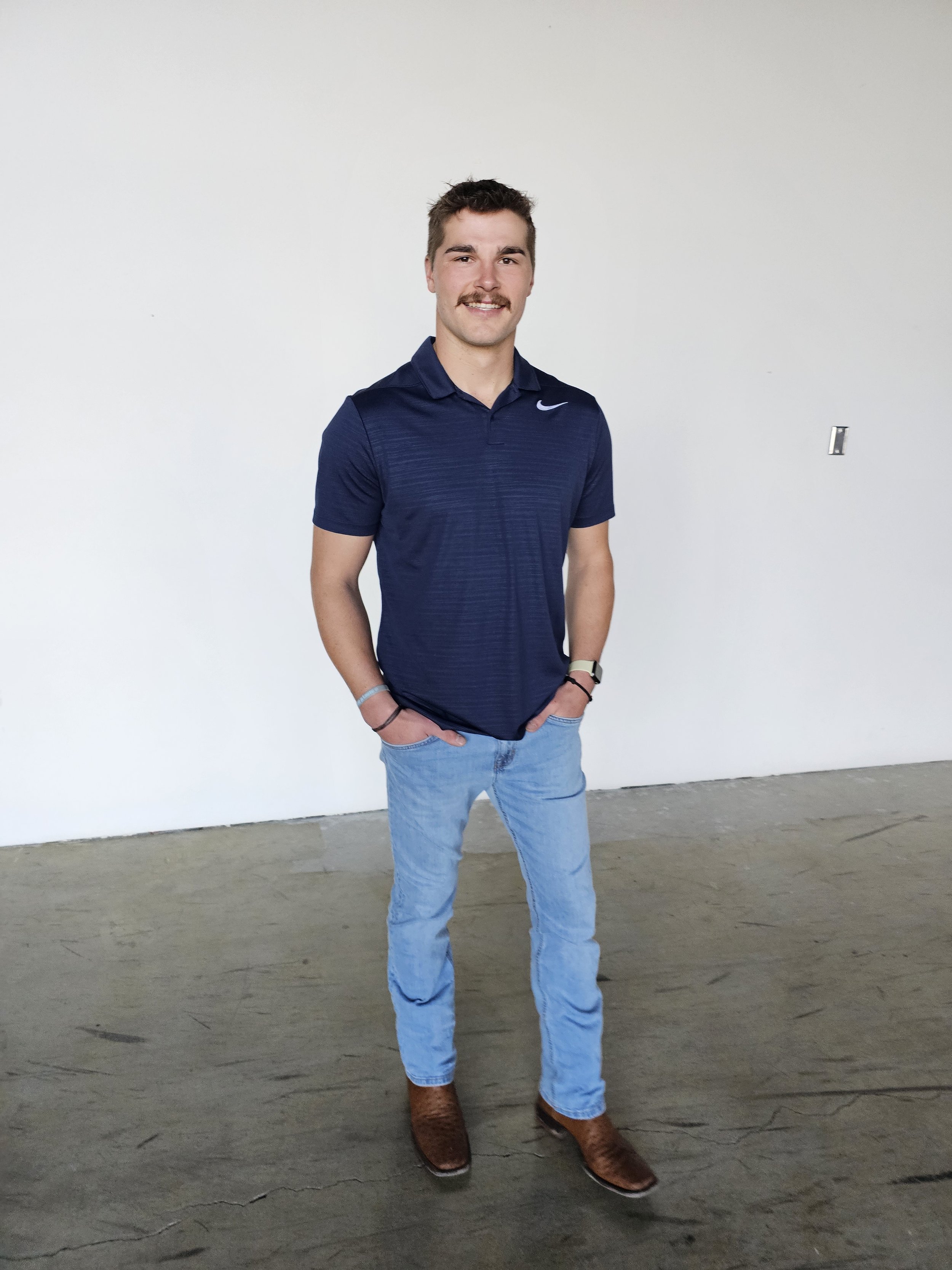 A young man with short brown hair, mustache, and beard, wearing a navy blue Nike polo shirt, light blue jeans, and brown cowboy boots standing against a plain white wall with his hands in his pockets, smiling at the camera.