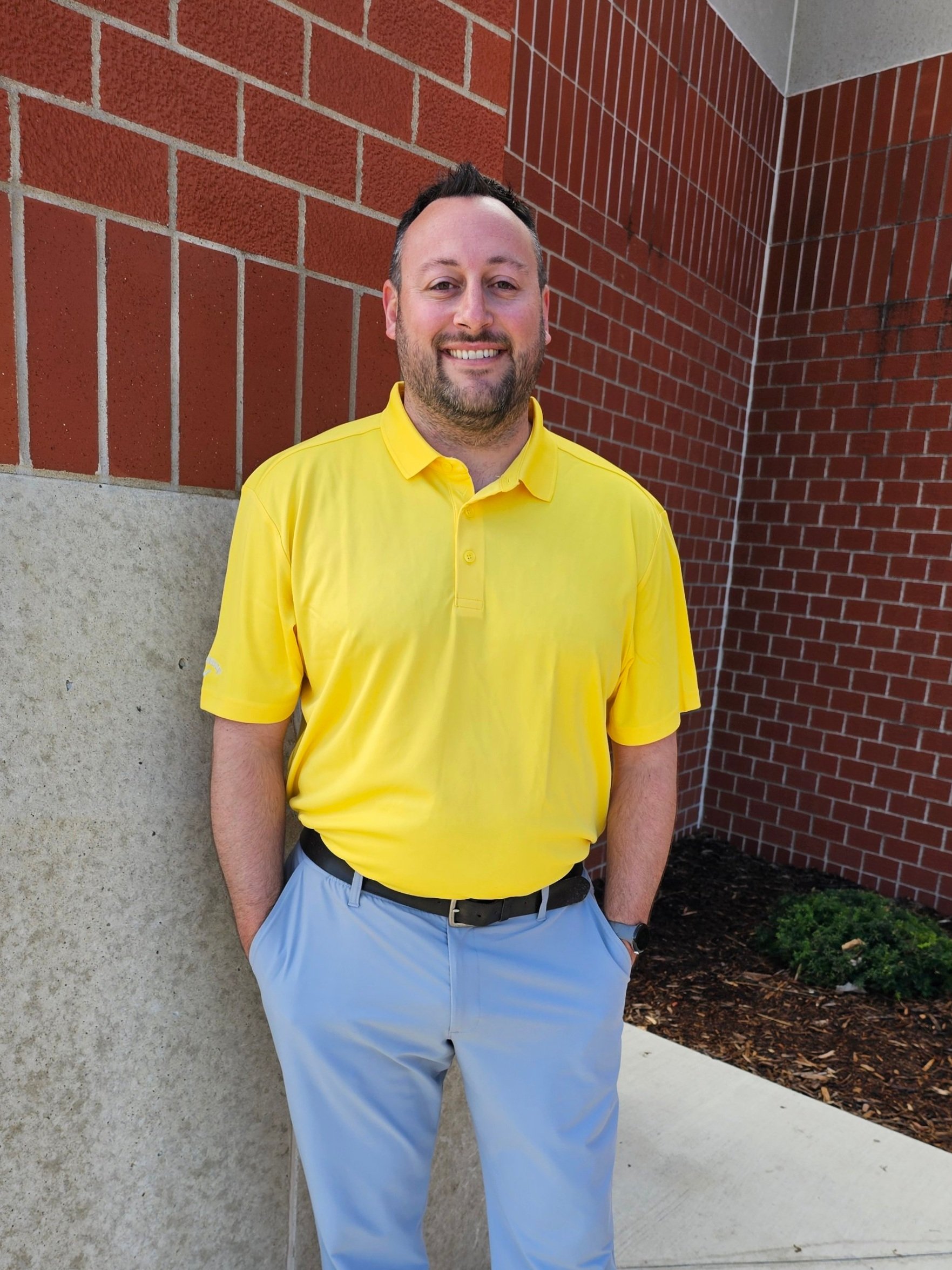 A man with a beard and short hair smiling, standing outdoors against a brick wall, wearing a yellow polo shirt and light gray pants.