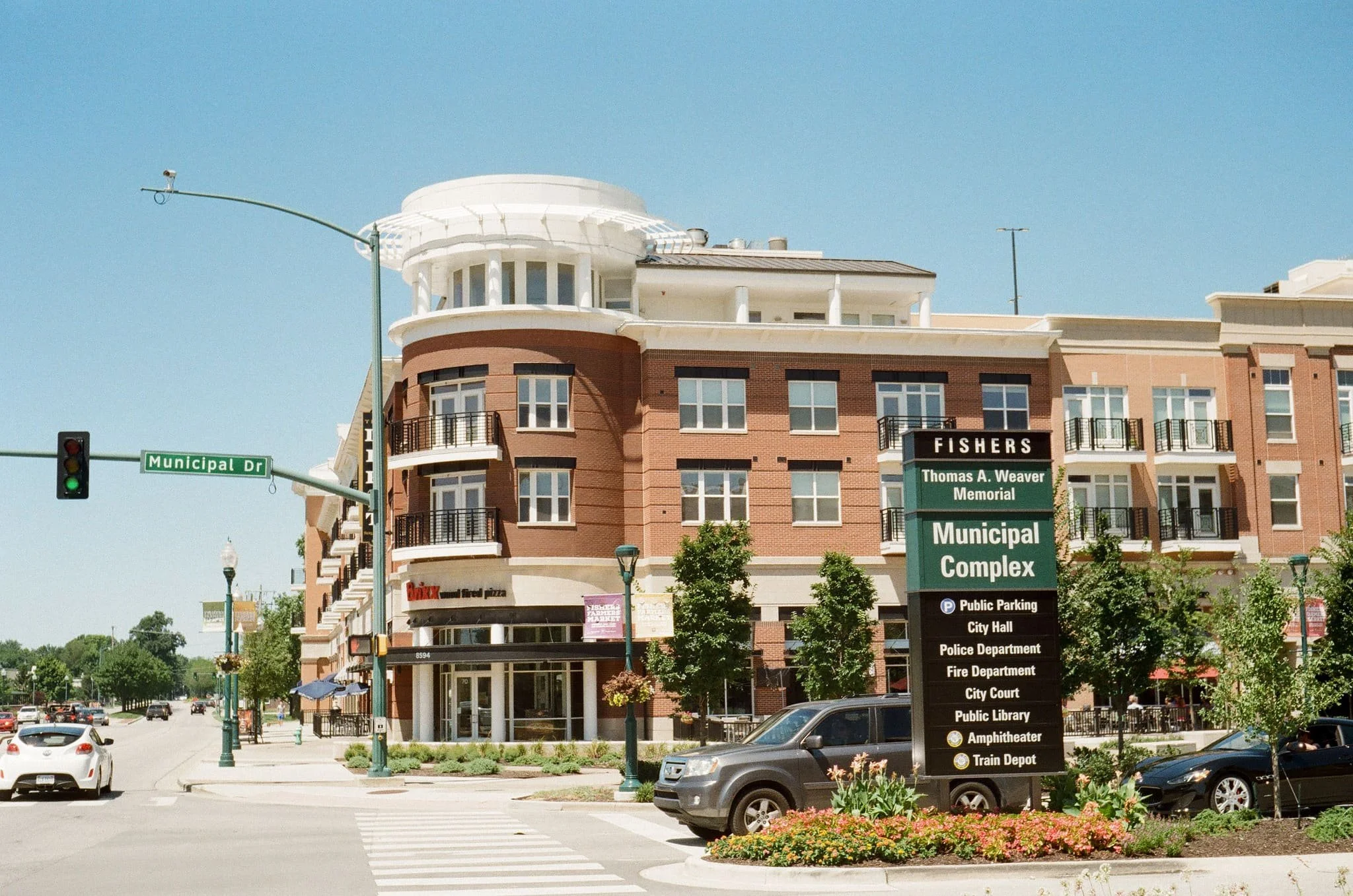 A multi-story building with brick and white exterior in an urban area with a street, cars, trees, and a large black and green sign listing public facilities and services.