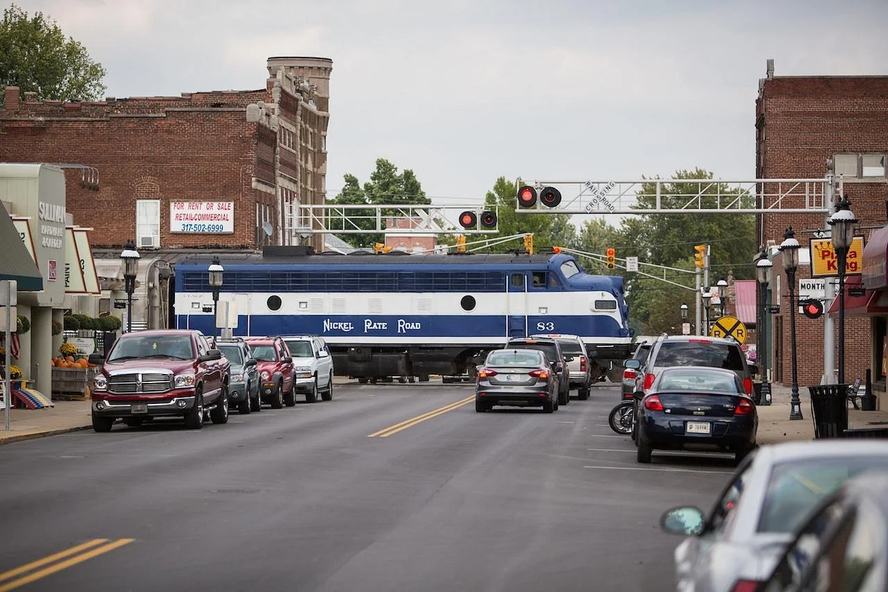 A blue Nickel Plate Road train passes through a small town's main street with cars parked on both sides. The street has buildings, street lamps, and traffic signals, and there are signs for a pizza place and rental spaces visible.