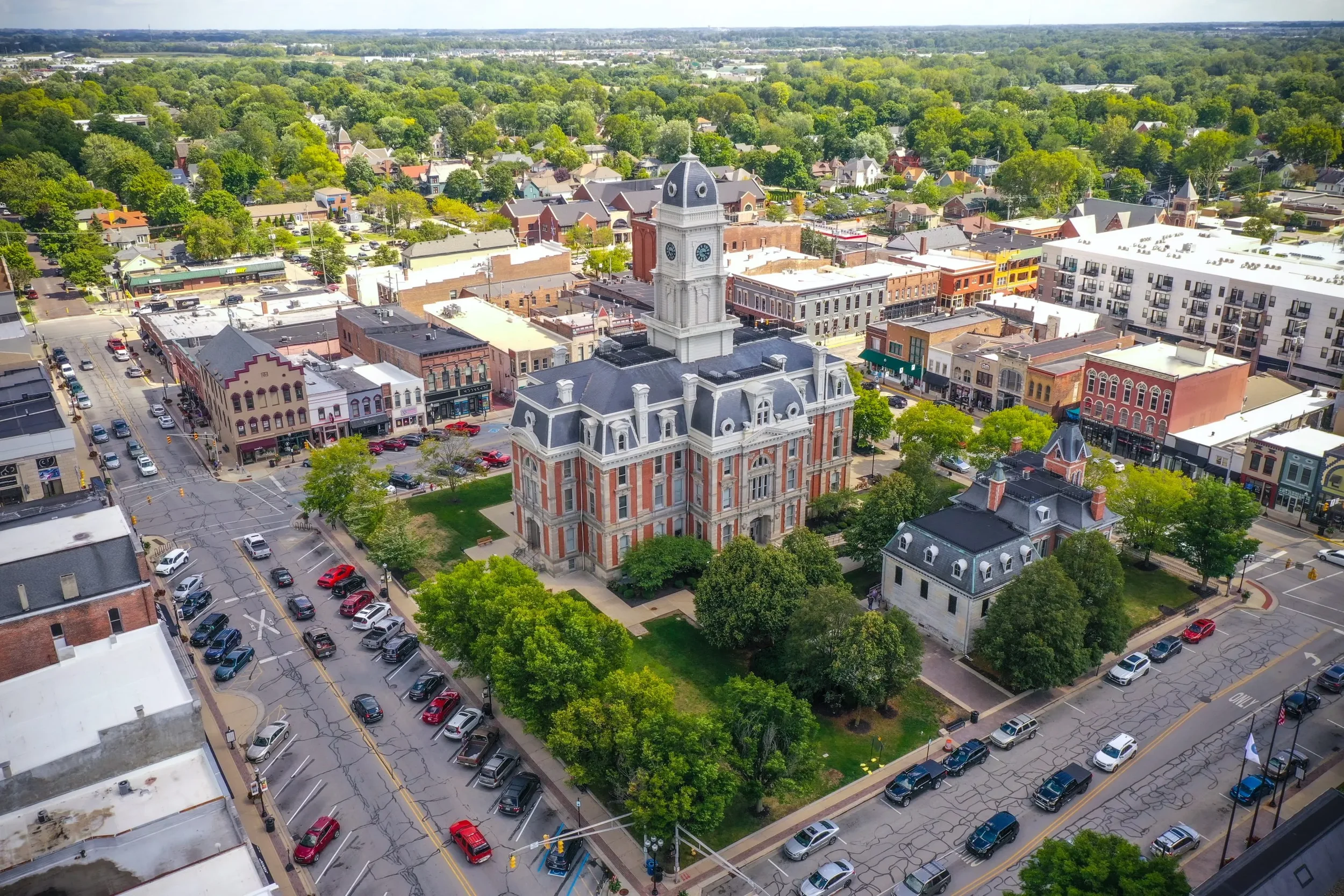 An aerial view of a historic courthouse with a tall clock tower, surrounded by a small park, located in a downtown area with various shops and buildings, with a mix of trees, parking lots, and a residential neighborhood in the background.