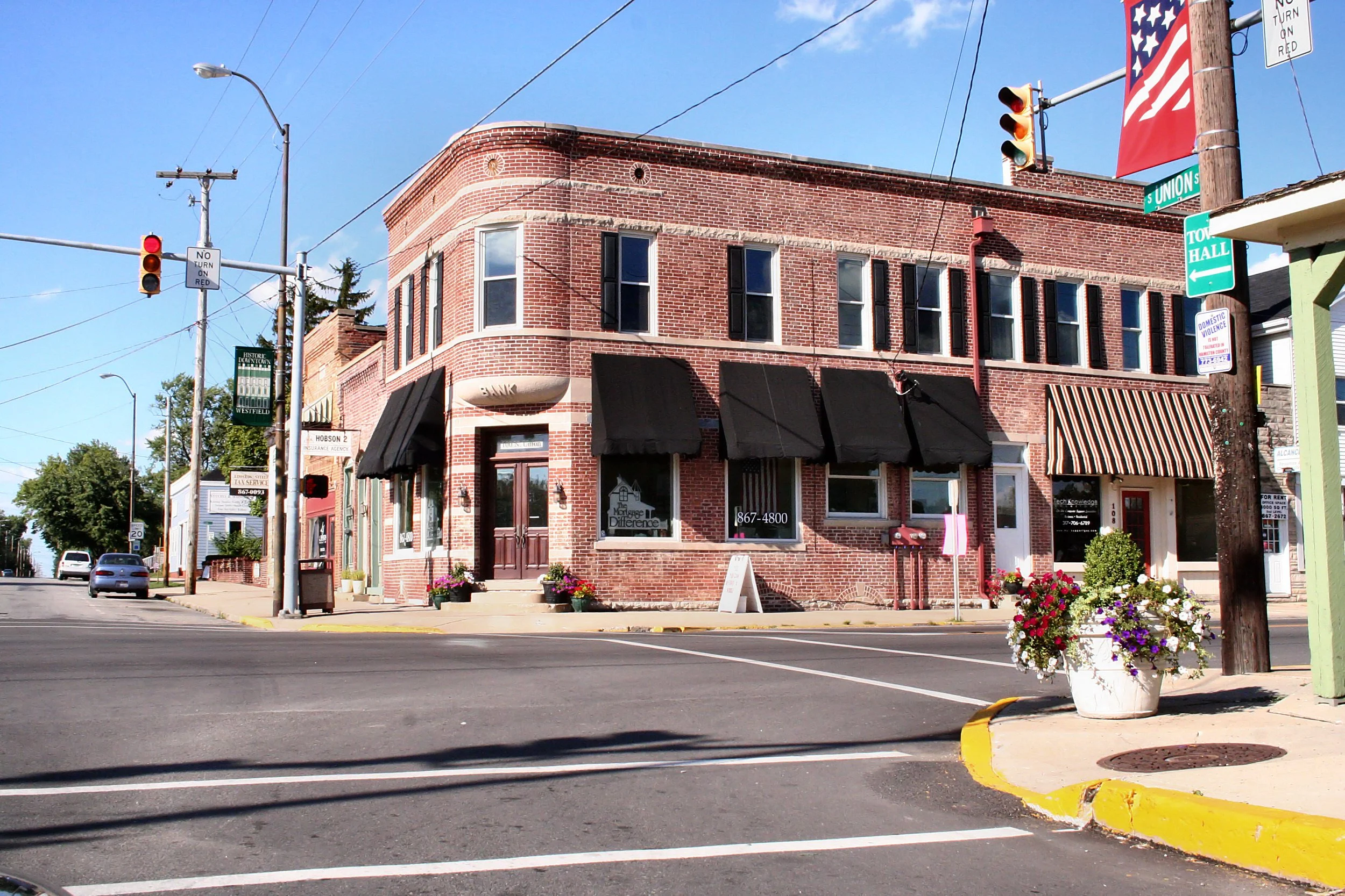 Street corner with brick buildings, traffic lights, street signs, flowers, and parked cars on a sunny day.