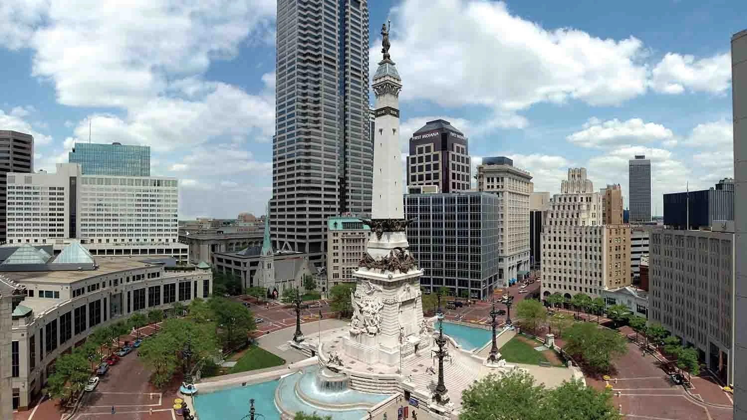 Downtown Indianapolis with monument square, a white memorial with statue at the top, surrounded by buildings, trees, and fountains, under a partly cloudy sky.