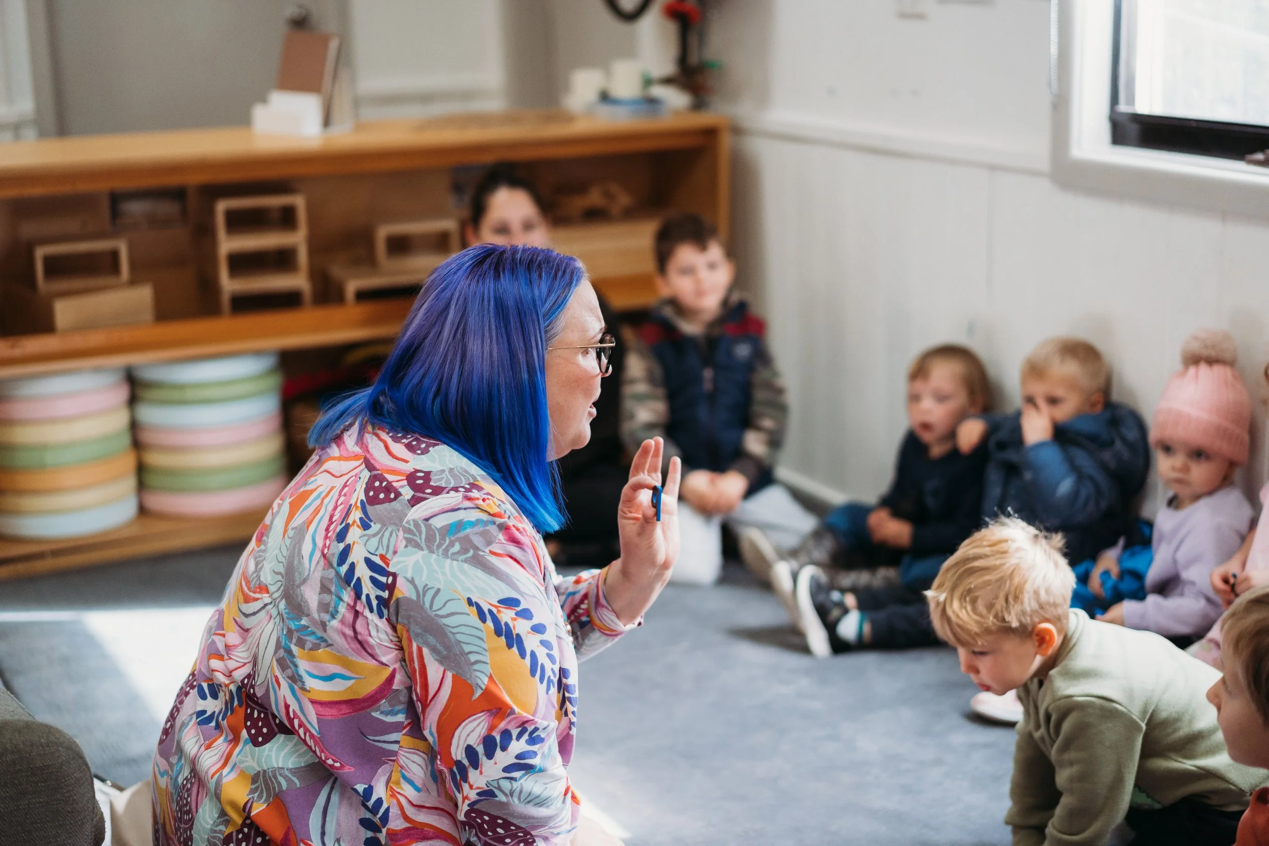 A woman with blue hair and glasses leading a group of children in a classroom or activity setting.