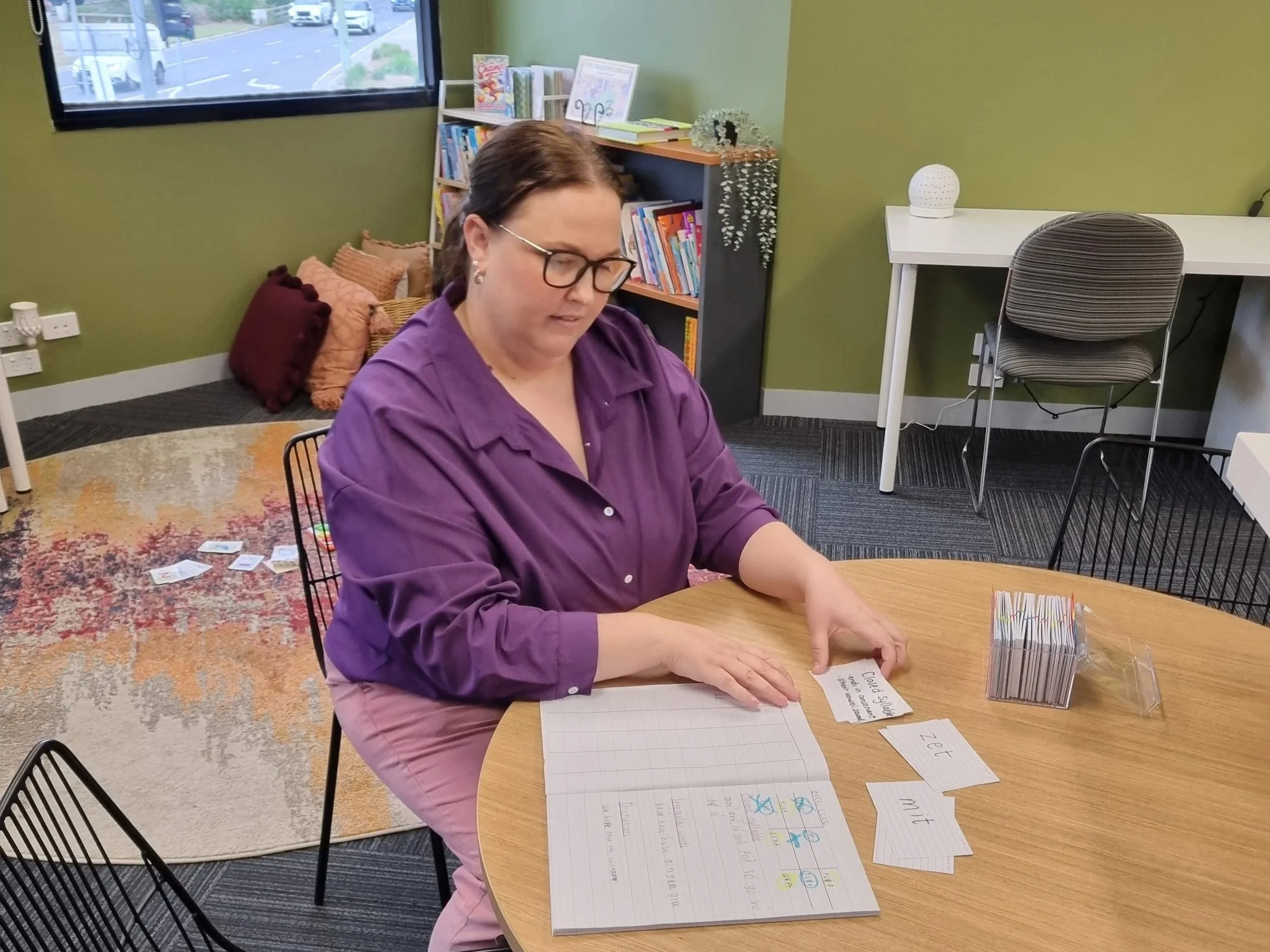 Woman with glasses wearing purple shirt sitting at a wooden table, working on a project with worksheets, paper, and markers, in a room with green walls, bookshelf, and a window showing cars outside.
