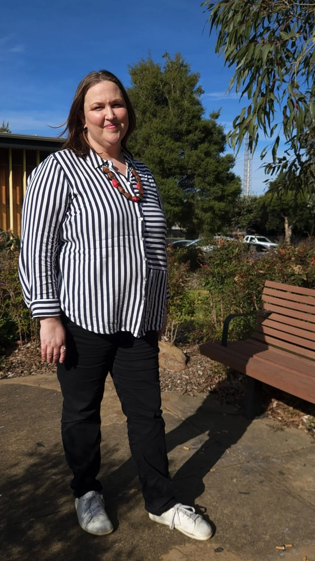 A woman wearing a black and white striped shirt, black pants, and white sneakers standing outdoors on a sunny day with trees and a wooden bench behind her.