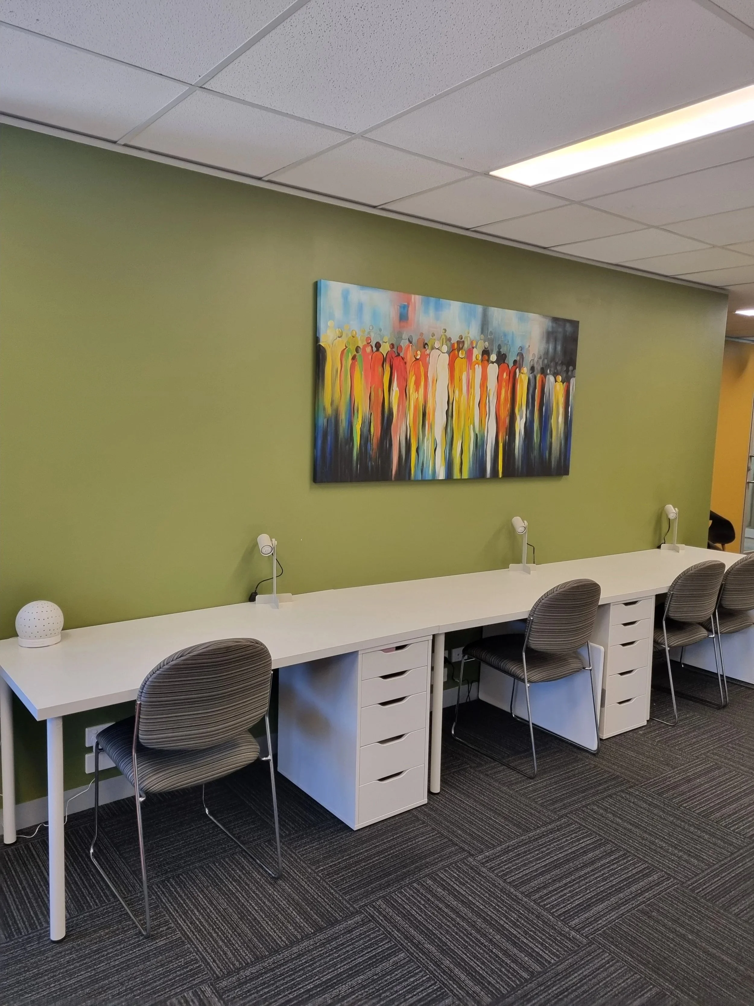 Three white desks with cushioned chairs tucked into them. A green wall with a colourful painting in the background.