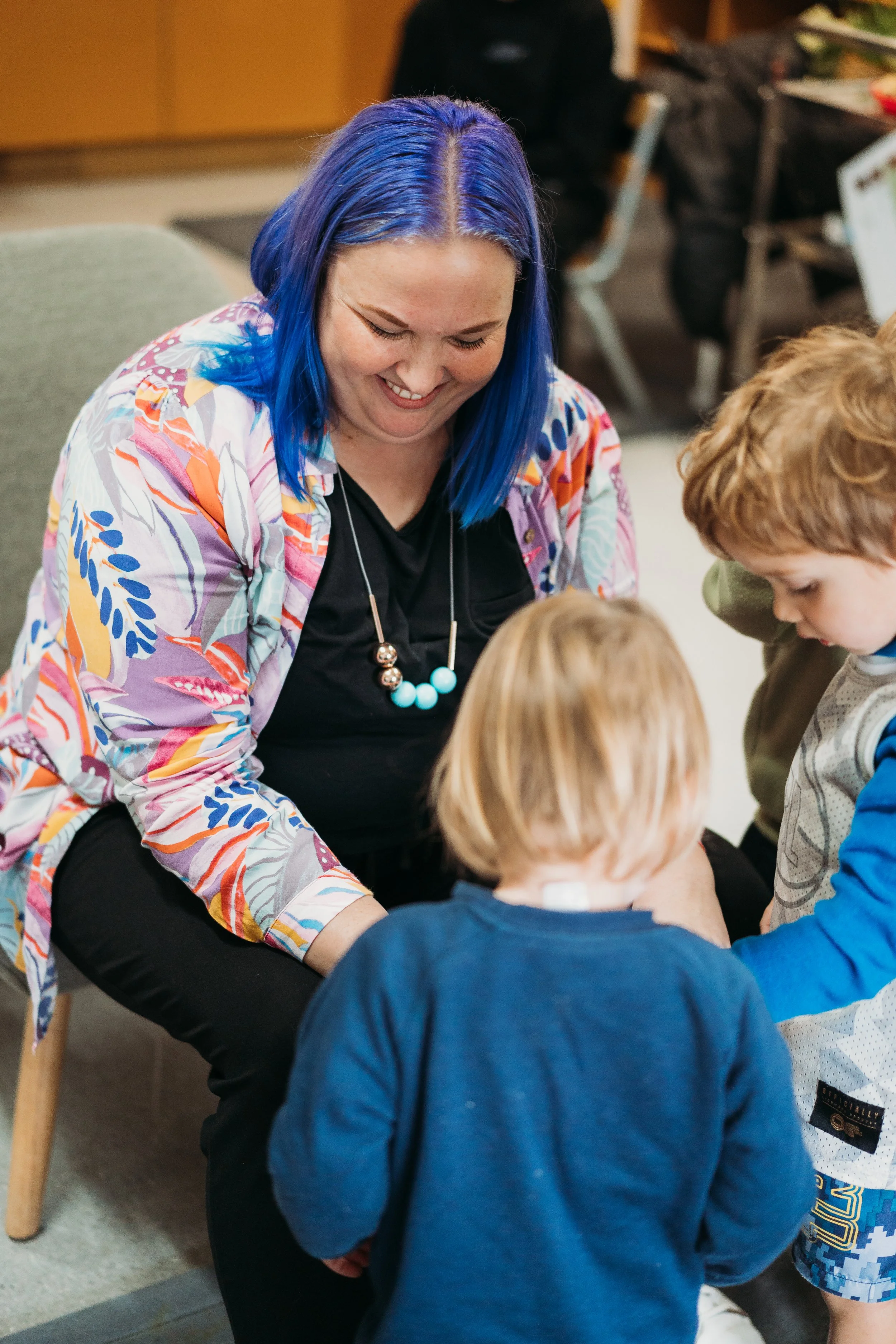 A woman with blue hair smiling while engaging with several children in a classroom setting.