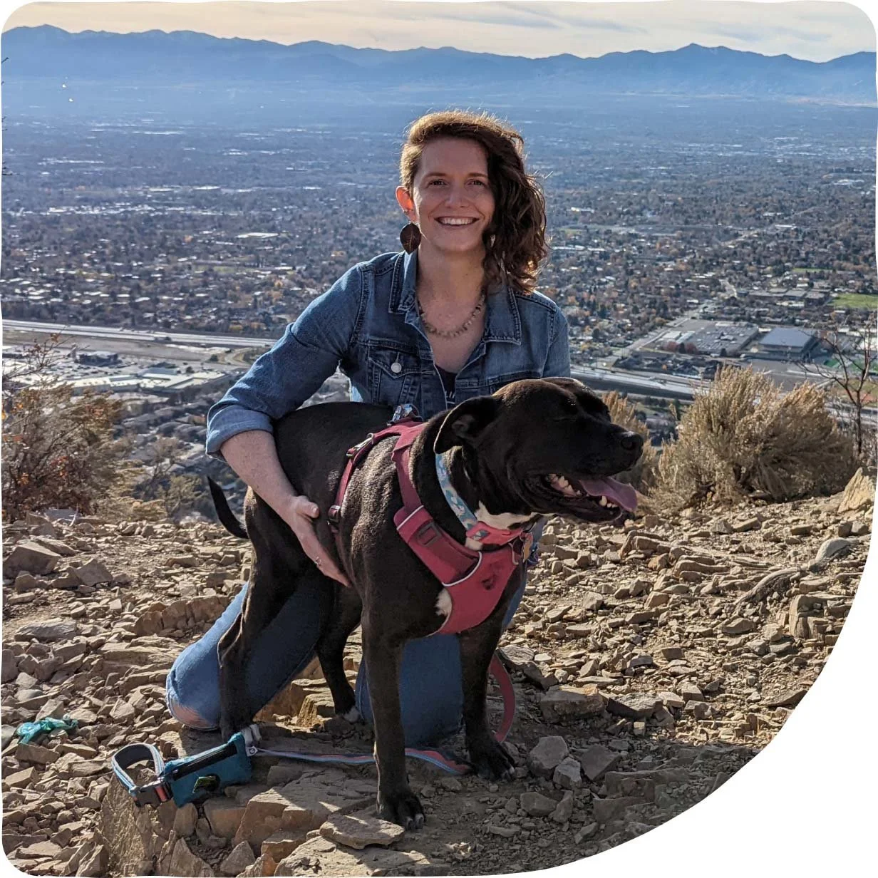 Alysha rose, a registered yoga teacher in salt lake city Utah, pictured on a sunny day in the mountains overlooking the Salt Lake Valley. Alysha is kneeling and hugging her dog in the photo.