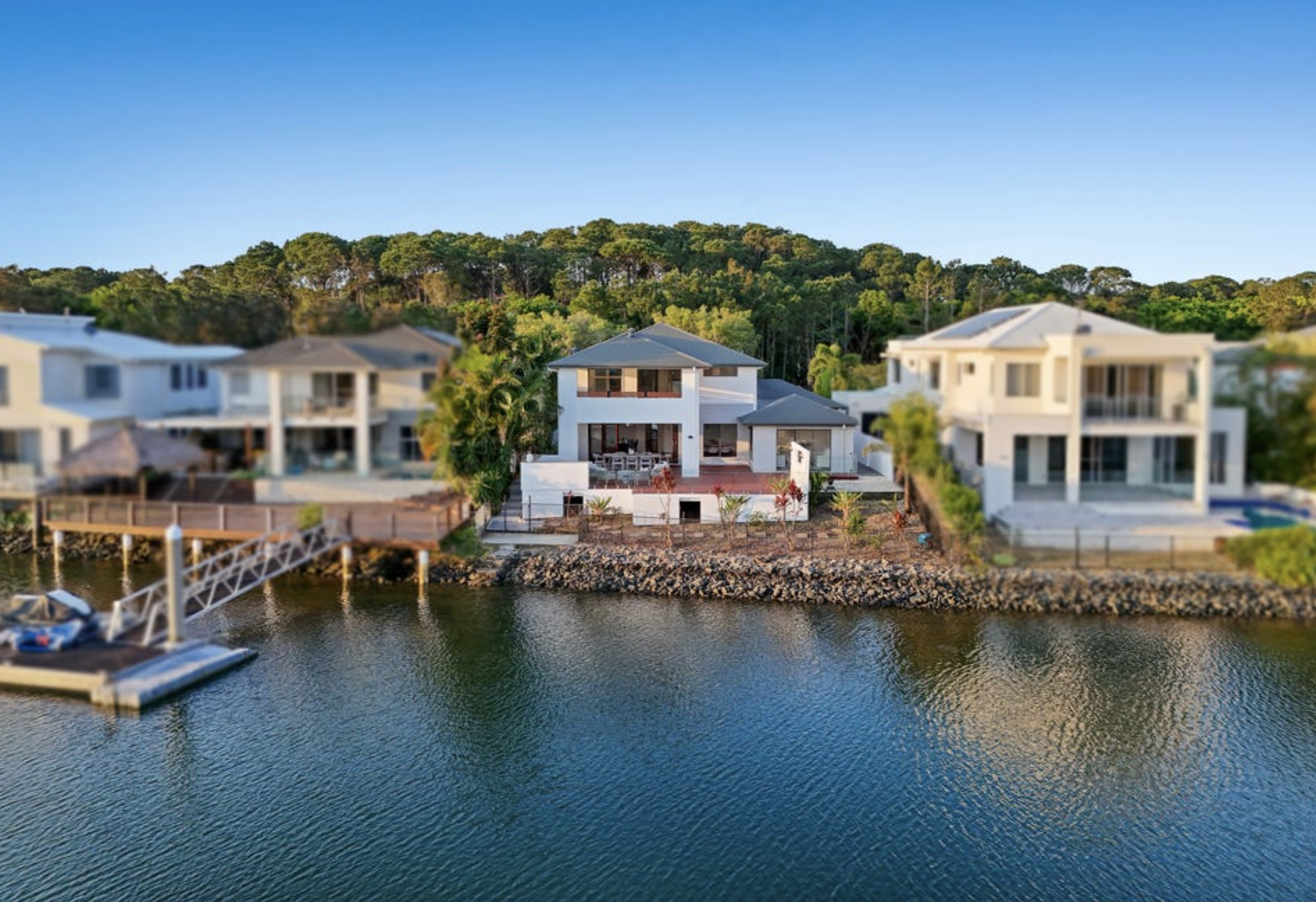 A waterfront home with a deck and above-ground pool, situated along a river with a dock and boat. Behind the house is a wooded hill with trees, under a clear blue sky.