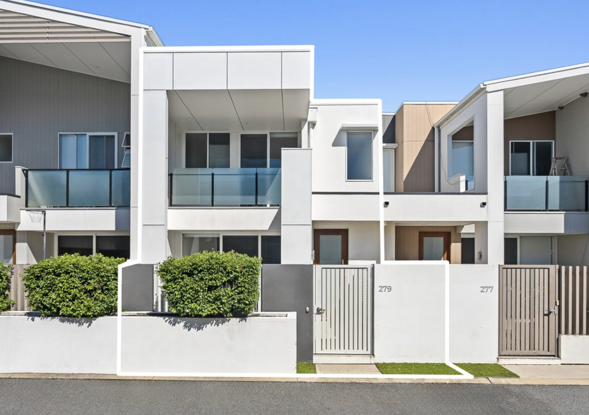 Modern multi-story residential buildings with balconies and glass railings, yellow and gray exteriors, surrounded by a white fence and greenery, under a clear blue sky.