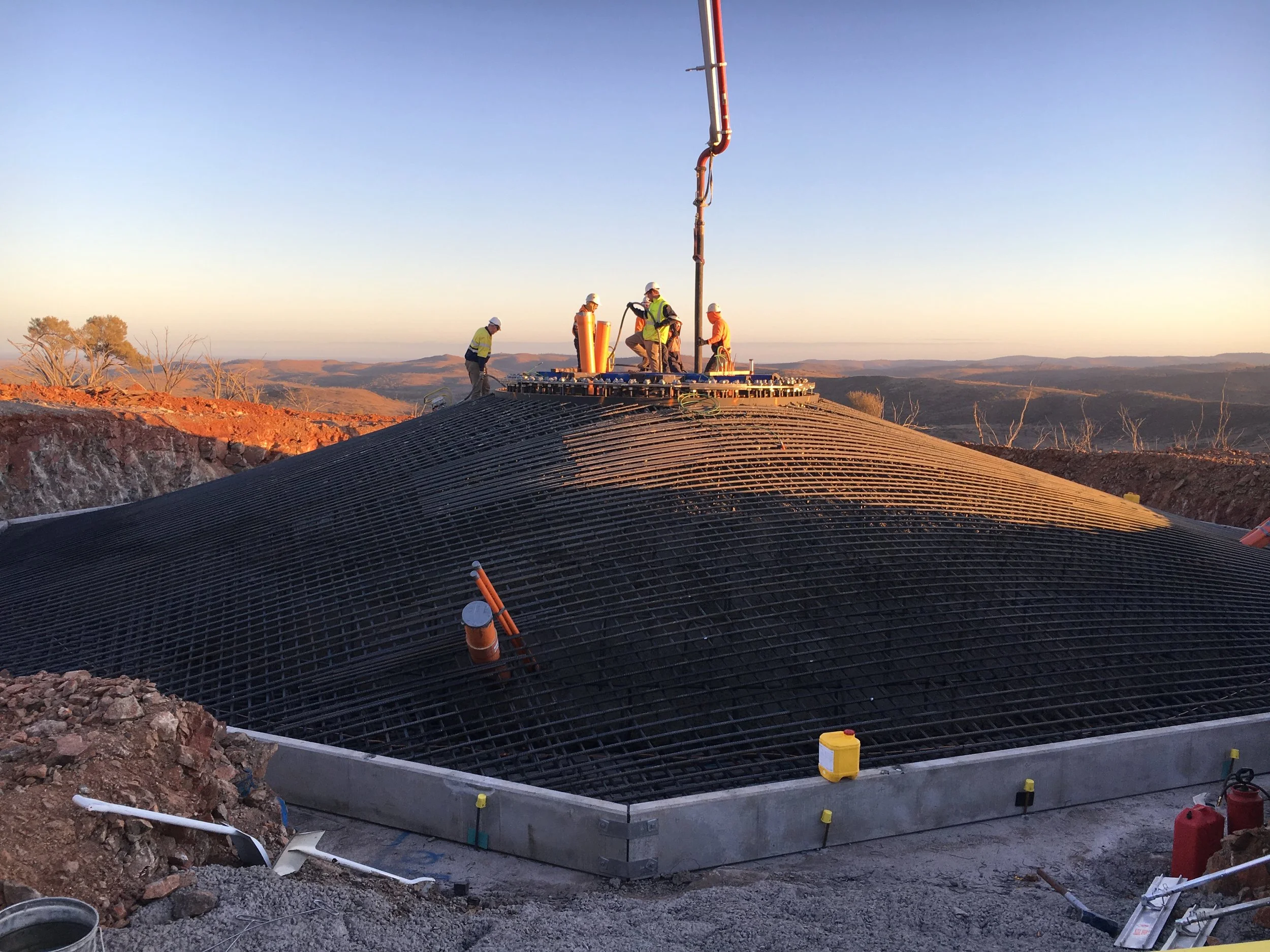 Construction workers installing a large underground water tank or similar structure with rebar framework on a hillside during sunset.