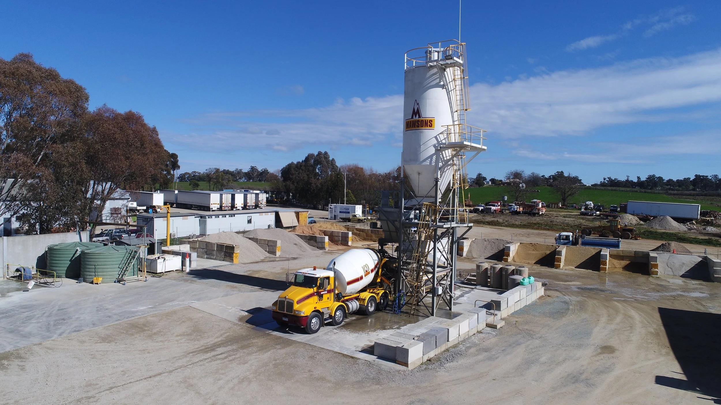 Construction site with a cement mixer truck and a tall cement silo marked 'Mawsons' under a blue sky, surrounded by dirt and construction materials.