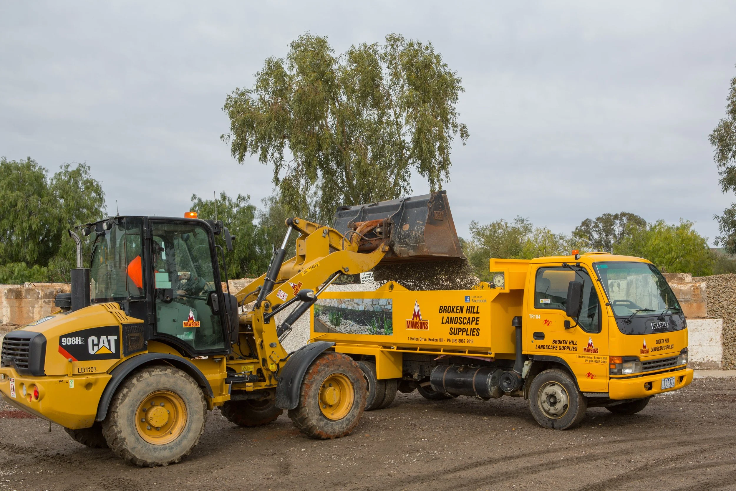 A yellow Caterpillar front loader dumping dirt into a yellow landscape maintenance truck marked with the logo of Broken Hill Landscape Supplies, parked on a construction site with trees in the background.