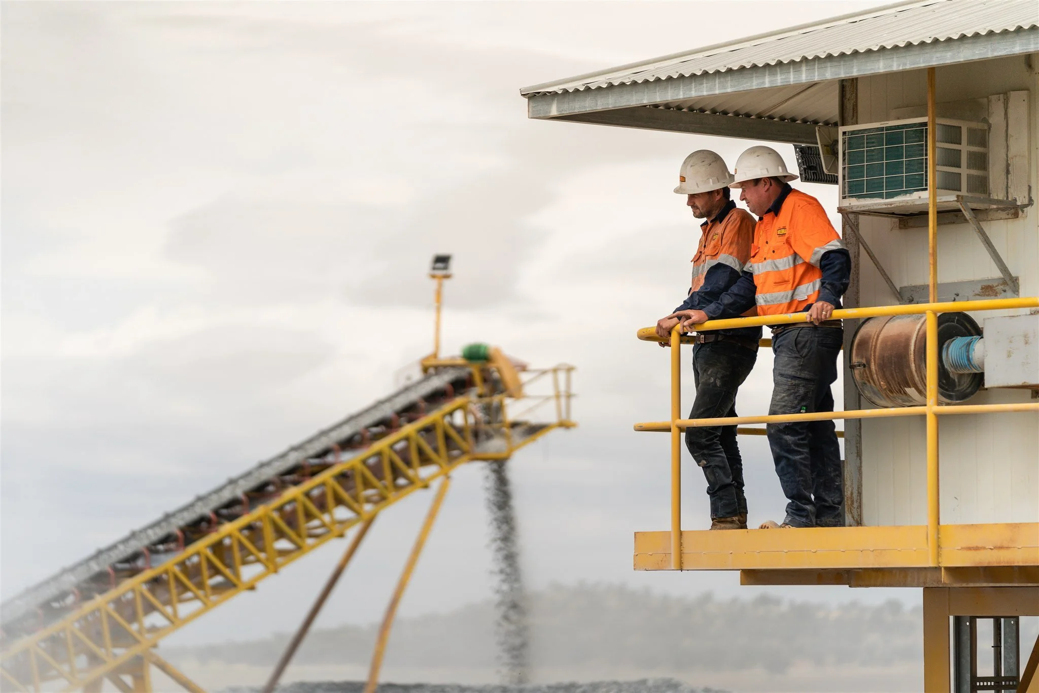 Two construction workers in orange safety vests and white helmets standing on a yellow platform, looking down at a conveyor system releasing crushed rock into a large pile.