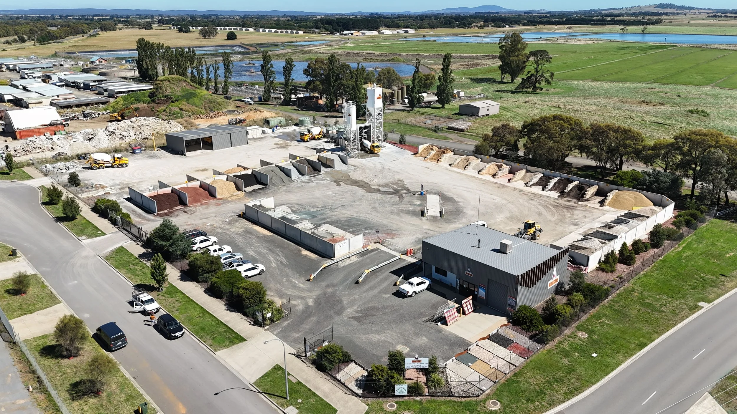 Aerial view of a construction and landscaping supply yard with piles of different colored soil and gravel, parked cars, and machinery, surrounded by fields and water bodies.