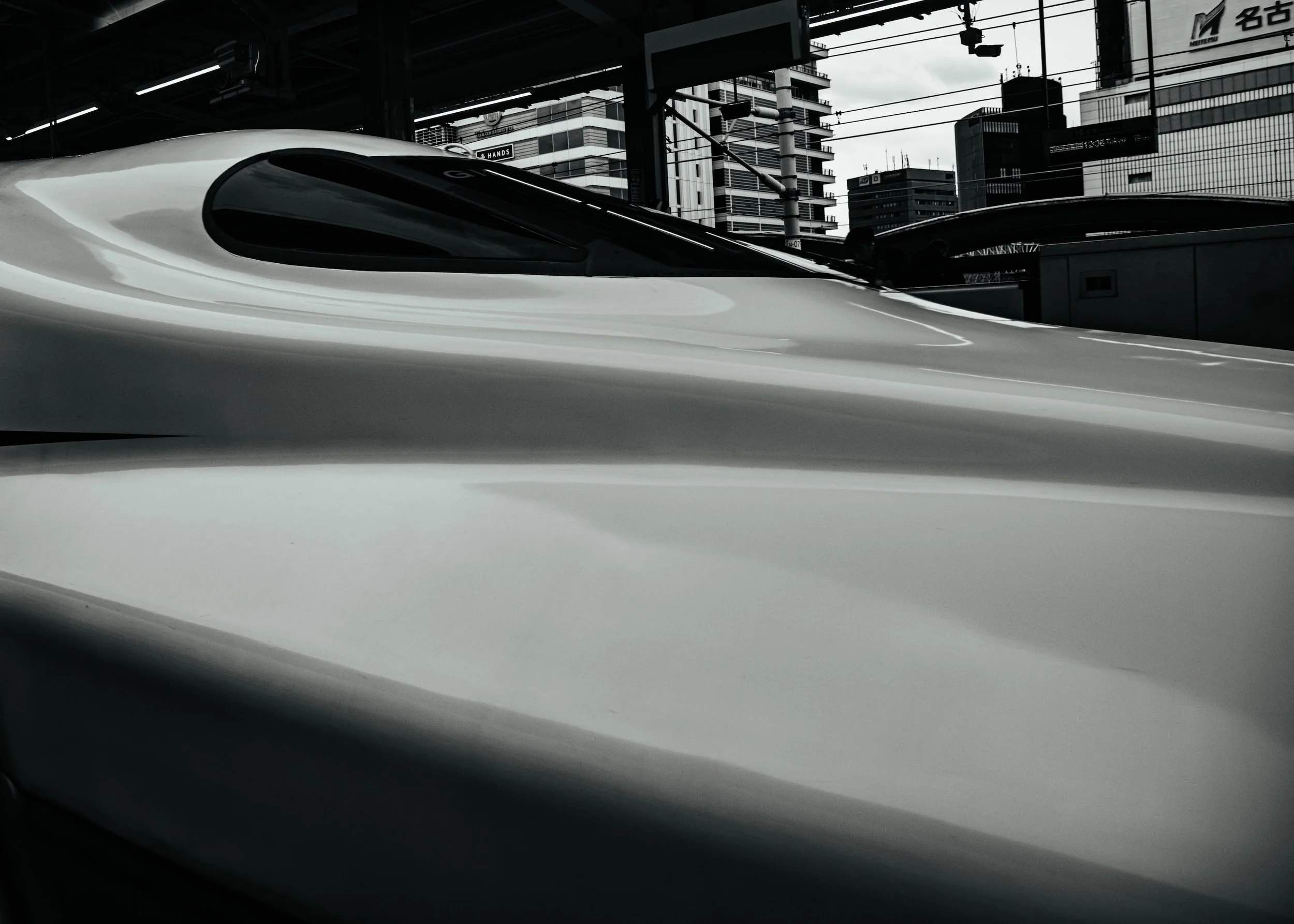 Black-and-white close-up of the sleek, elongated nose of a Shinkansen bullet train at a Nagoya station platform, its curved white bodywork filling the frame, city towers and station signage rising behind.