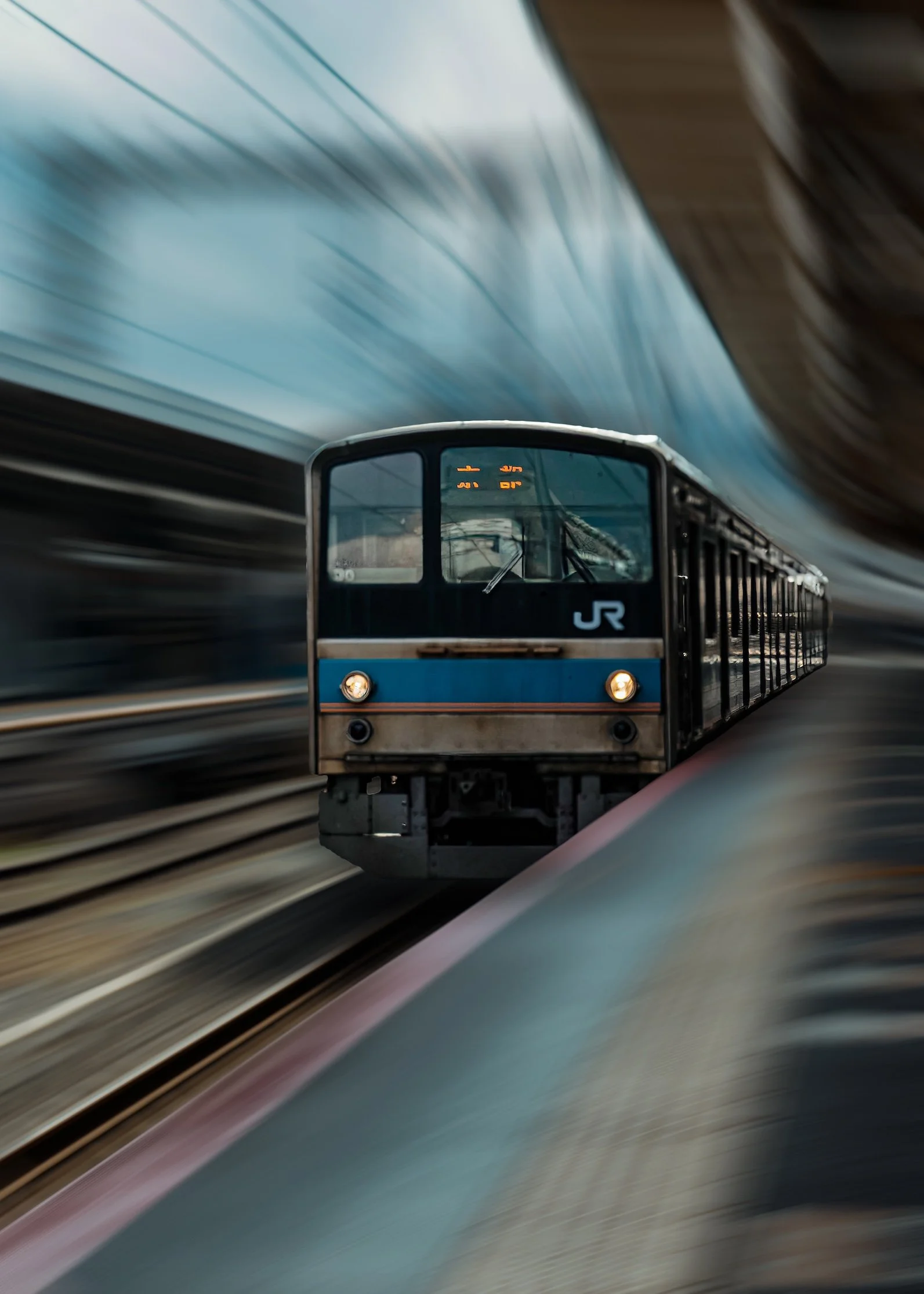 JR commuter train with a blue stripe and lit headlamps rushing head-on toward the camera, the whole surrounding scene stretched into radial motion blur that pulls the eye into the sharp, still front carriage.