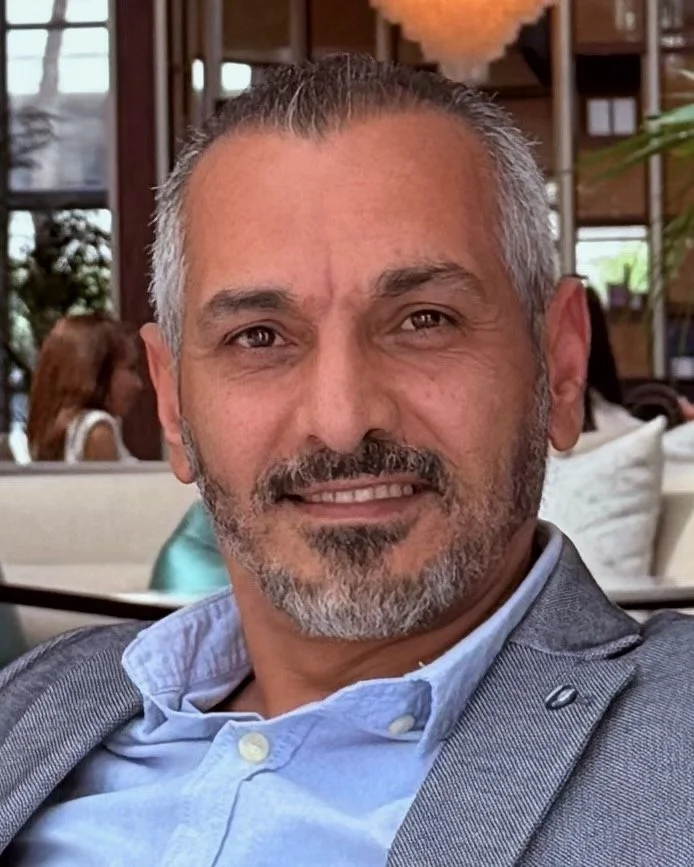 A middle-aged man with short gray hair and a beard, wearing a light blue collared shirt and a gray blazer, smiling at the camera in a well-lit restaurant or lounge setting.