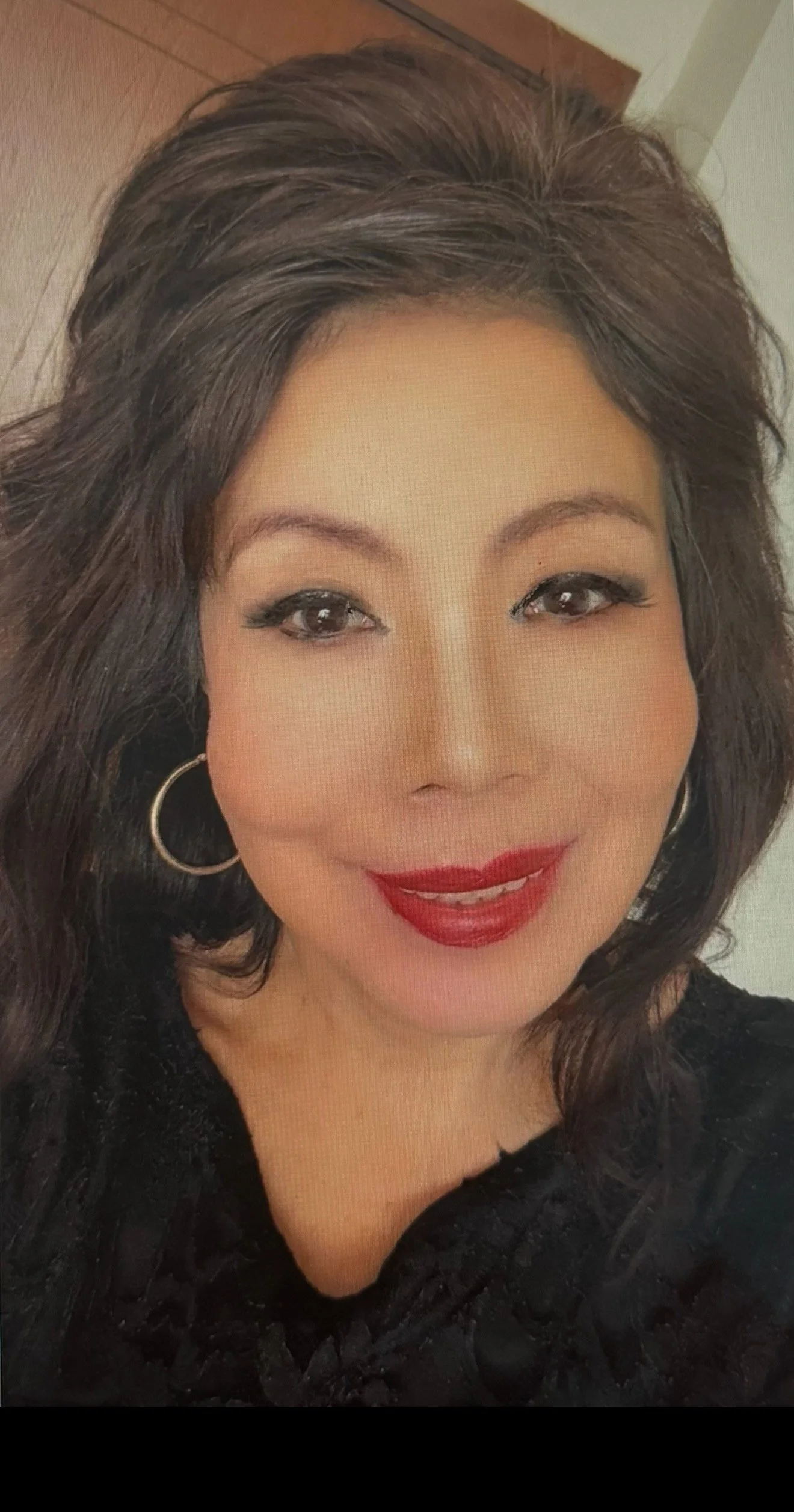 A woman with dark brown, shoulder-length hair smiling, wearing silver hoop earrings and red lipstick, dressed in a black top, indoors with a plain background.