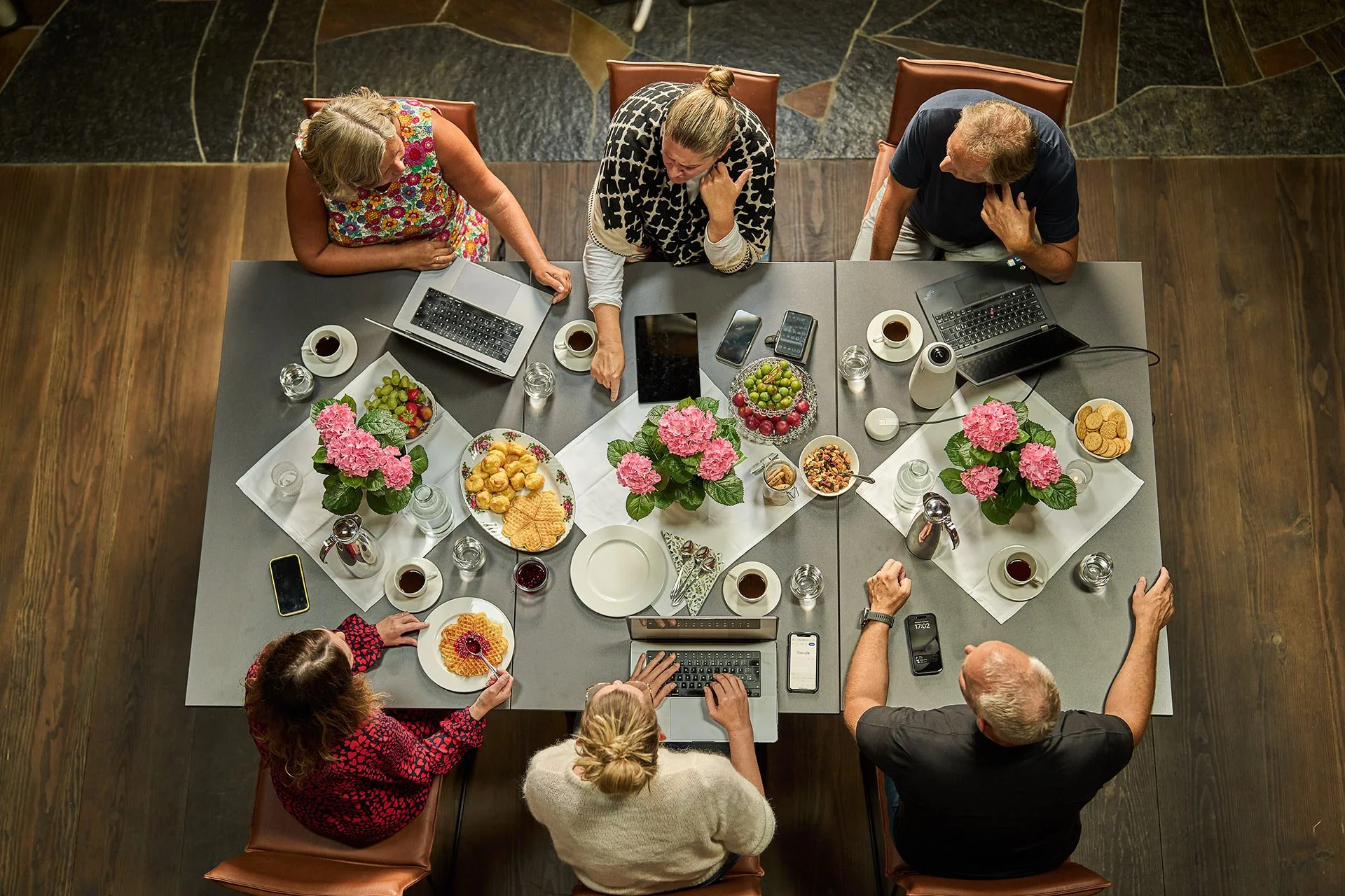Syv personer sitter rundt et bord med datamaskiner, telefoner og mat, sett ovenfra.