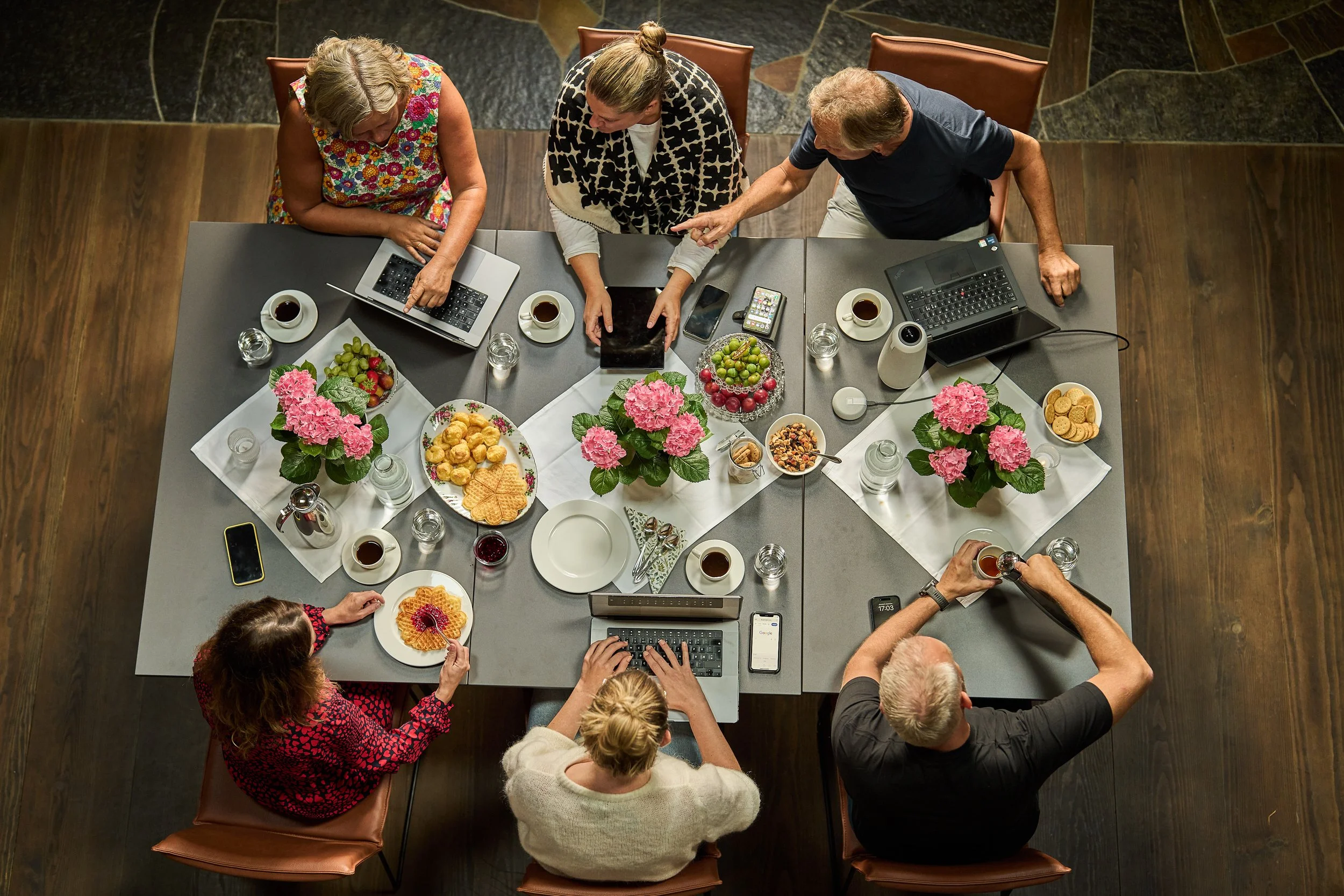 Seks personer sitter rundt et bord med flasker, kaffekopper, frukt og kaker. De bruker bærbare datamaskiner og telefoner. Fra et fugleperspektiv.