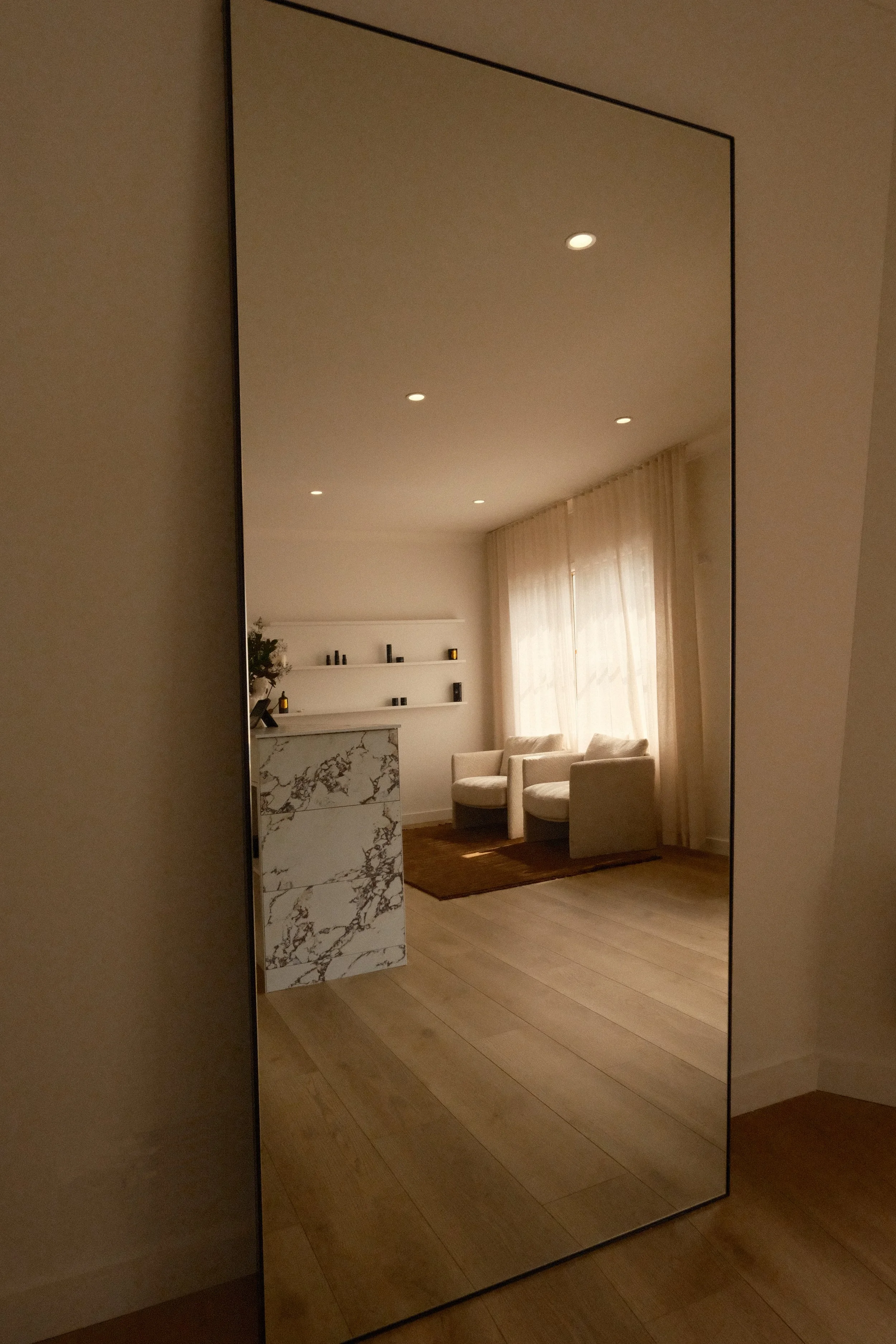 Reflection of a living room with two white chairs, a brown rug, curtains, and floating shelves with decor.