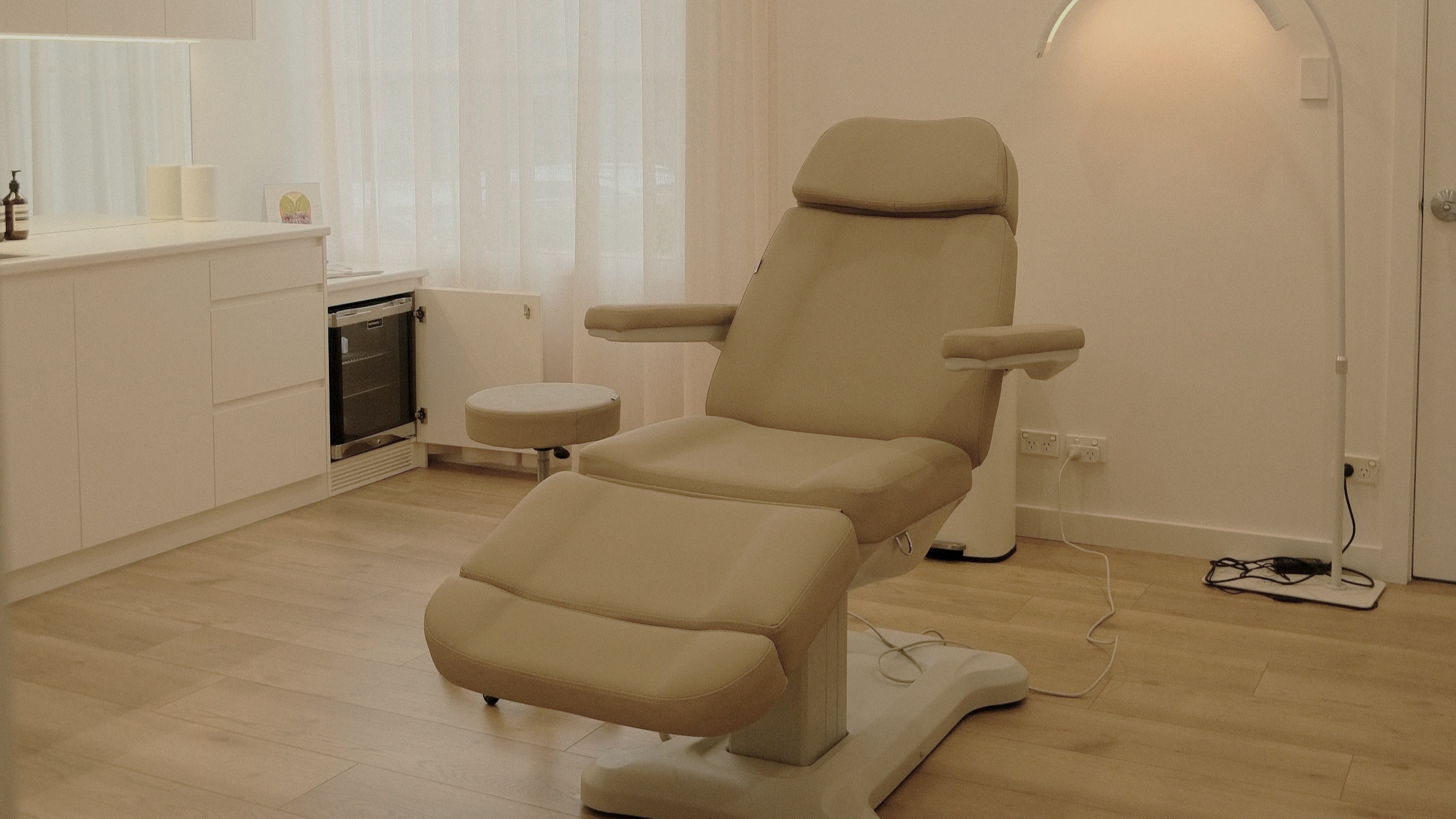 Medical examination room with beige reclining chair, small stool, white cabinet, and floor lamp with curved stand.
